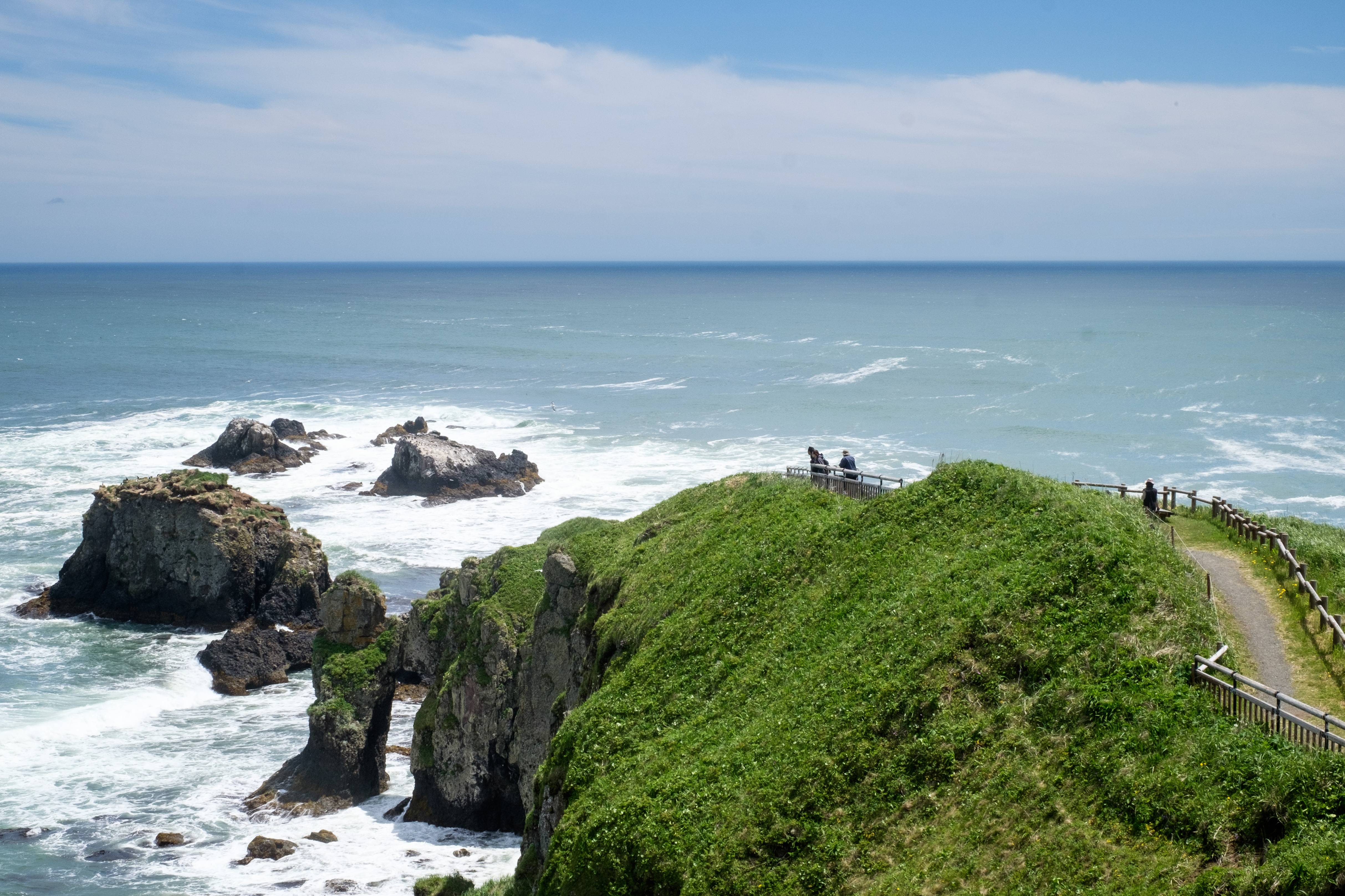A group of walkers make their way out to a wild headland. The cliffs are surrounded by white water as waves crash into shore.