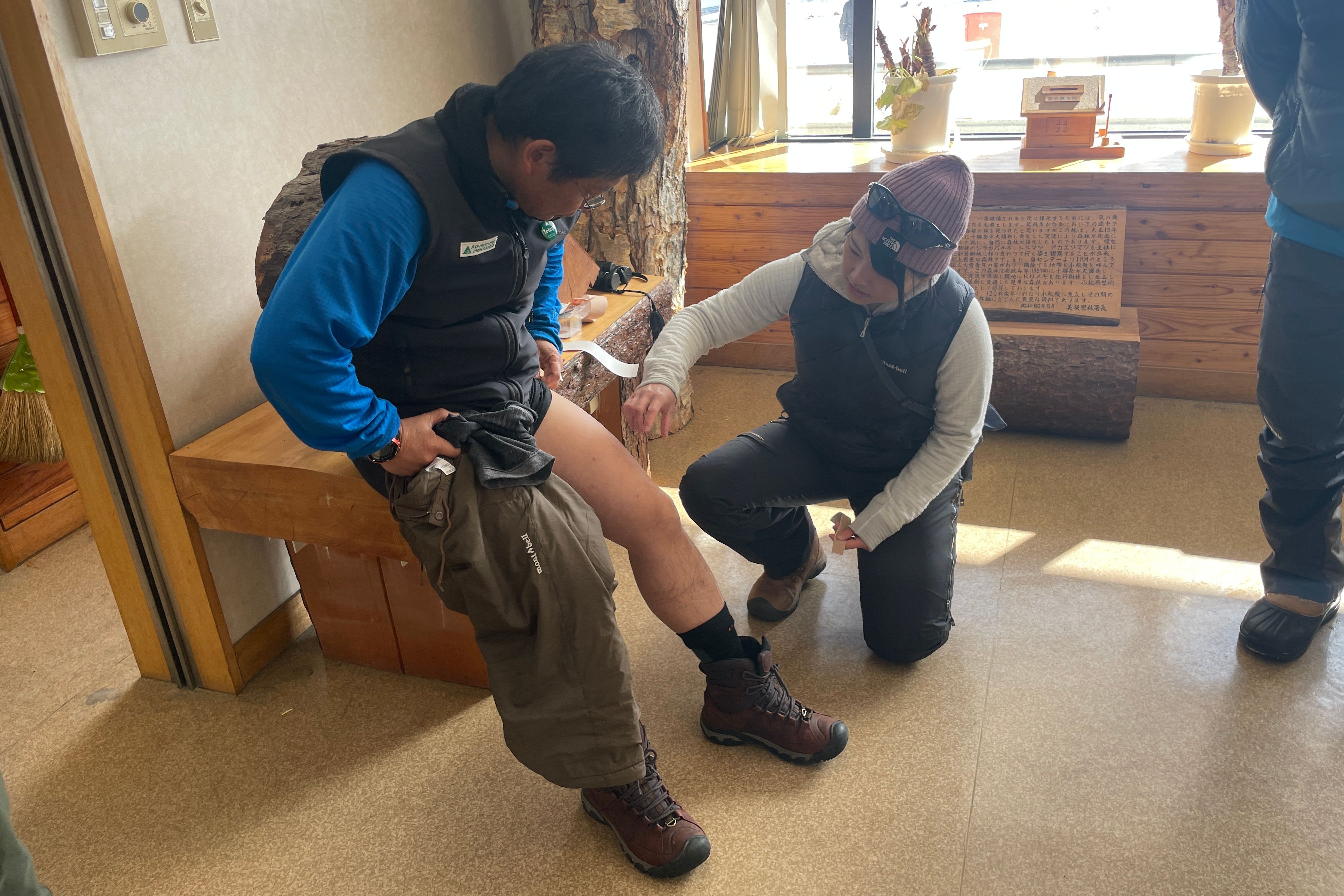 A hiking guide, who is also a physiotherapist, kneels to demonstrate how to apply therapeutic tape to another guide's knee for pain relief.