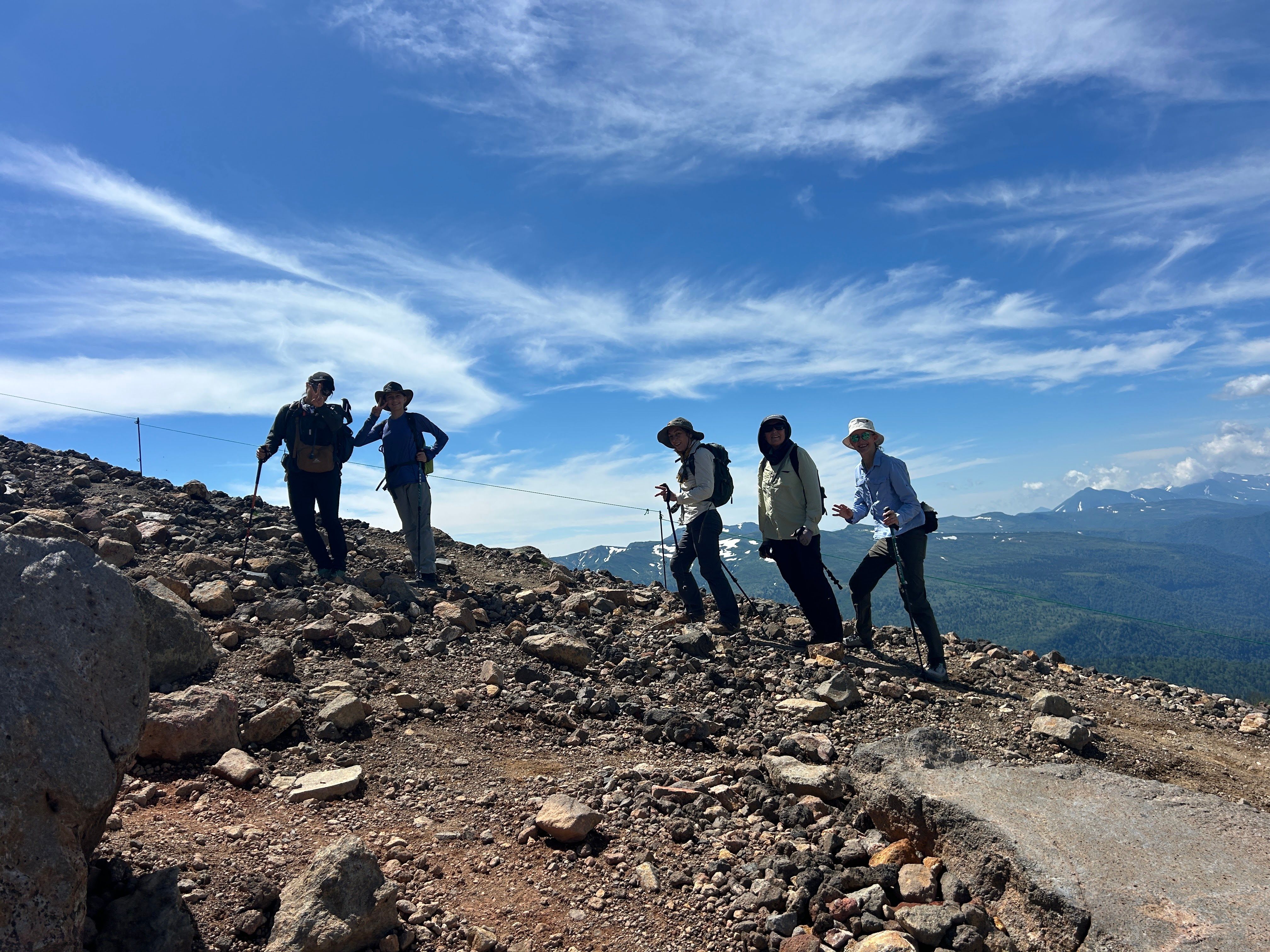 A group of hikers smile at the camera on a ridgeline on Mt. Asahidake. It is a very sunny day and there are cobweb-like clouds in the sky. Behind the hikers, Daisetsuzan National Park is clearly visible.