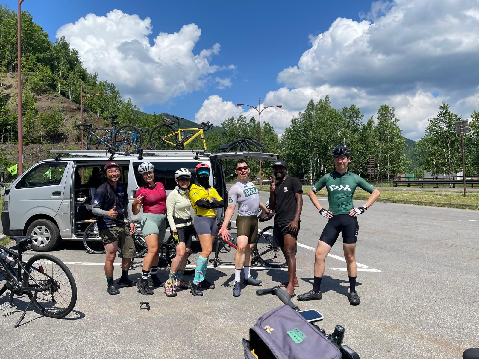 A group of cyclists pose in their cycling gear in front of a parked support van. Bicycles in various stages of assembly are around the parking lot.