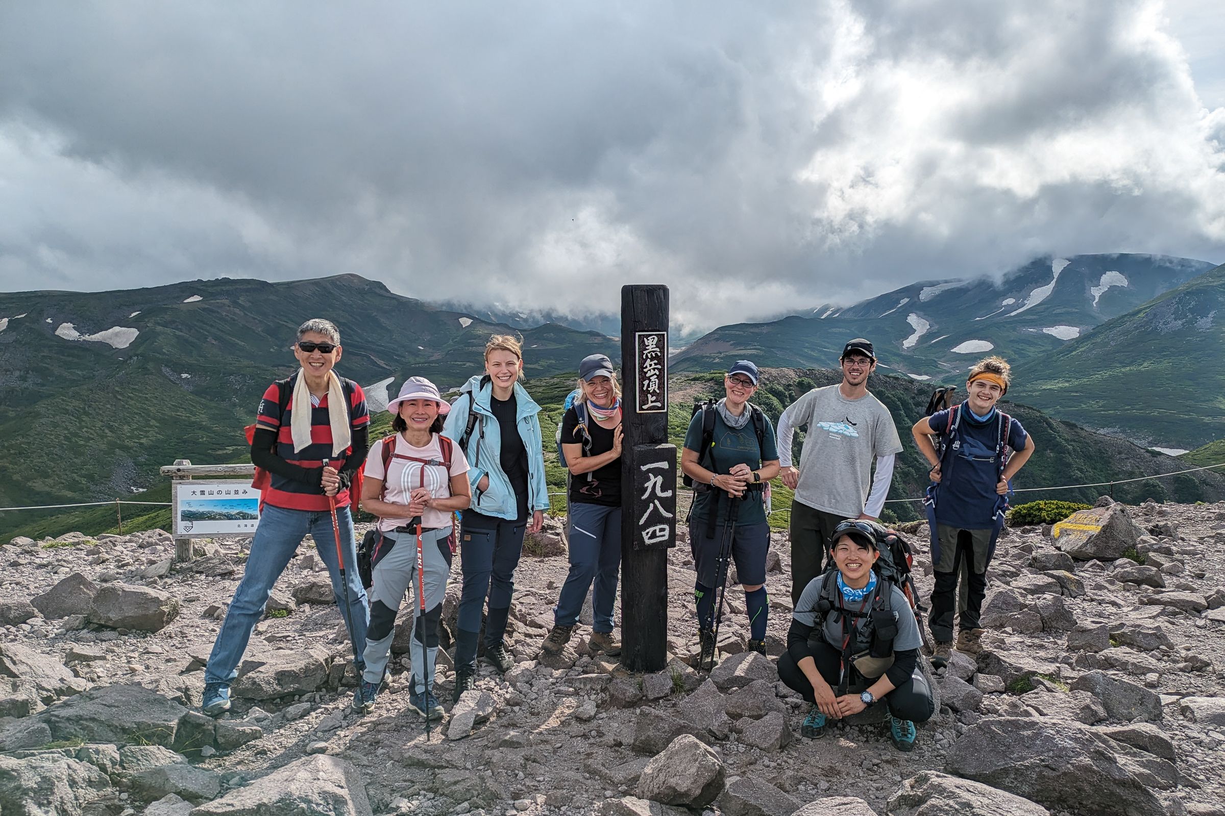 A group of eight hikers smile at the summit of a mountain. A pole denoting the summit in Japanese reads "Kurodake Summit - 1984m")