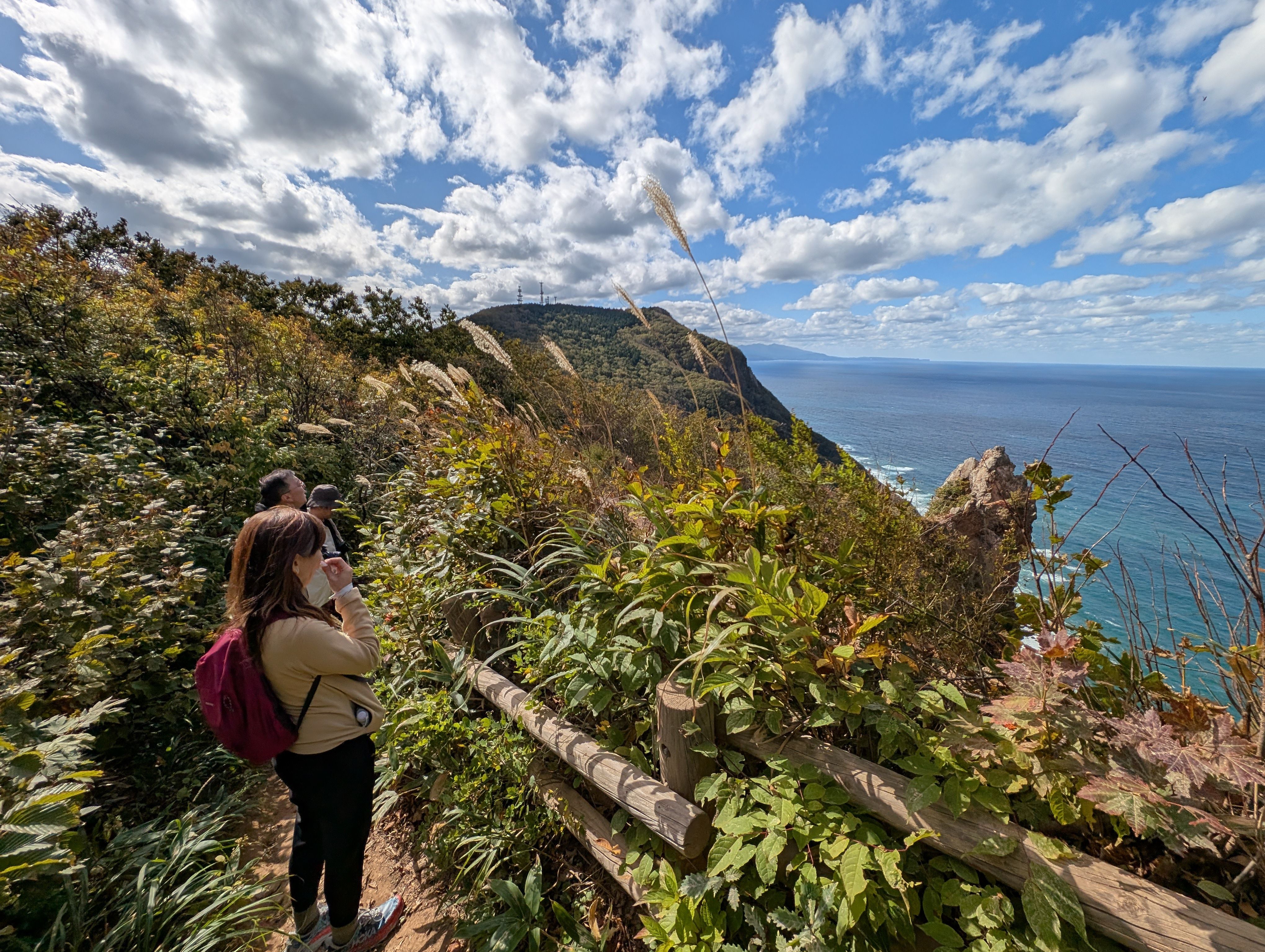 A line of hikers admire an ocean view visible from the clifftops of Mt. Akaiwa in Otaru. It is a sunny day. the sea and sky are both blue. Early autumn colours are visible on the clifftop.