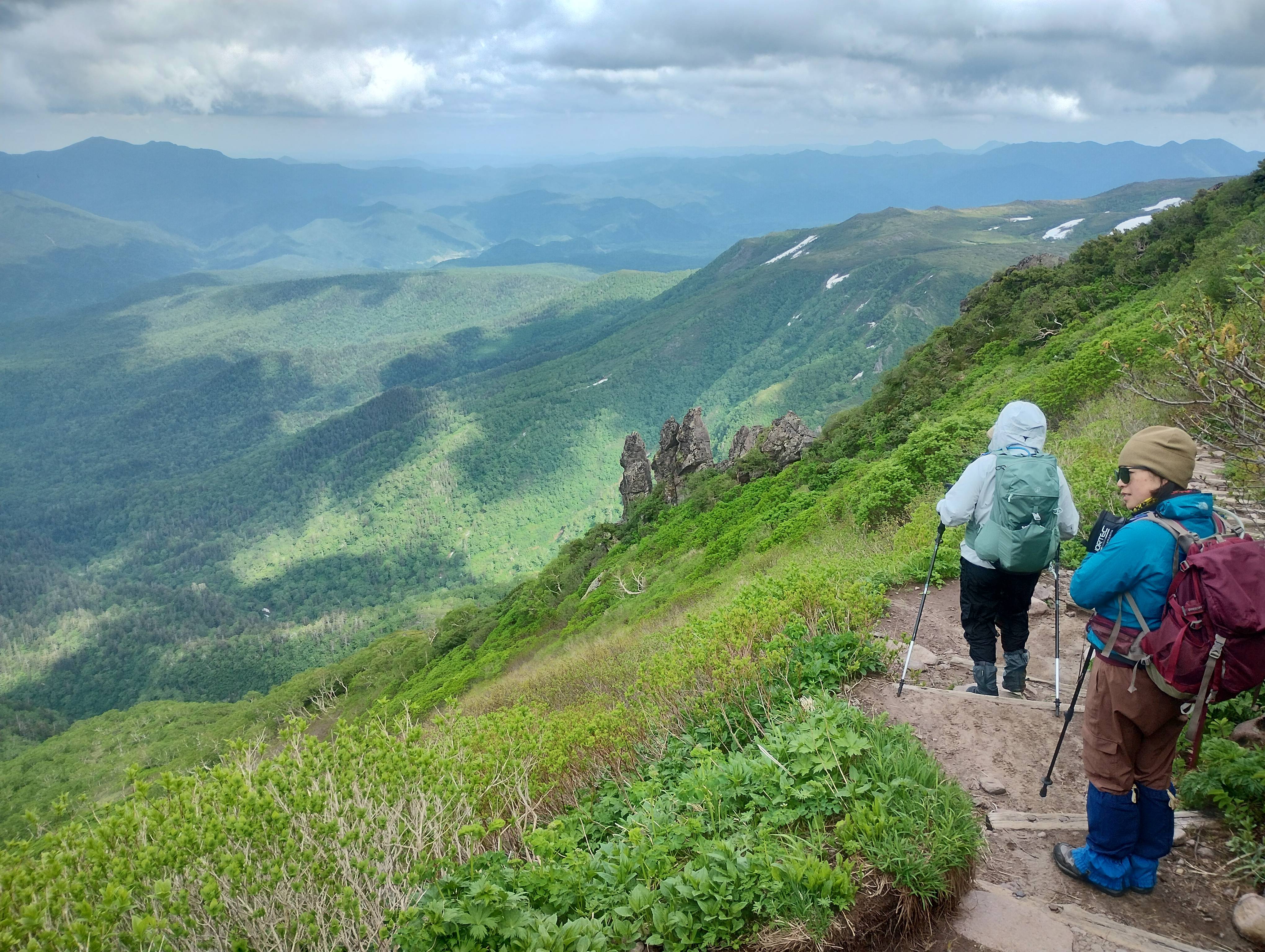 Two hikers descend the mountain path on Mt. Kurodake, Hokkaido. one of them looks out at a valley stretching below them, in which the shadows of clouds are visible.