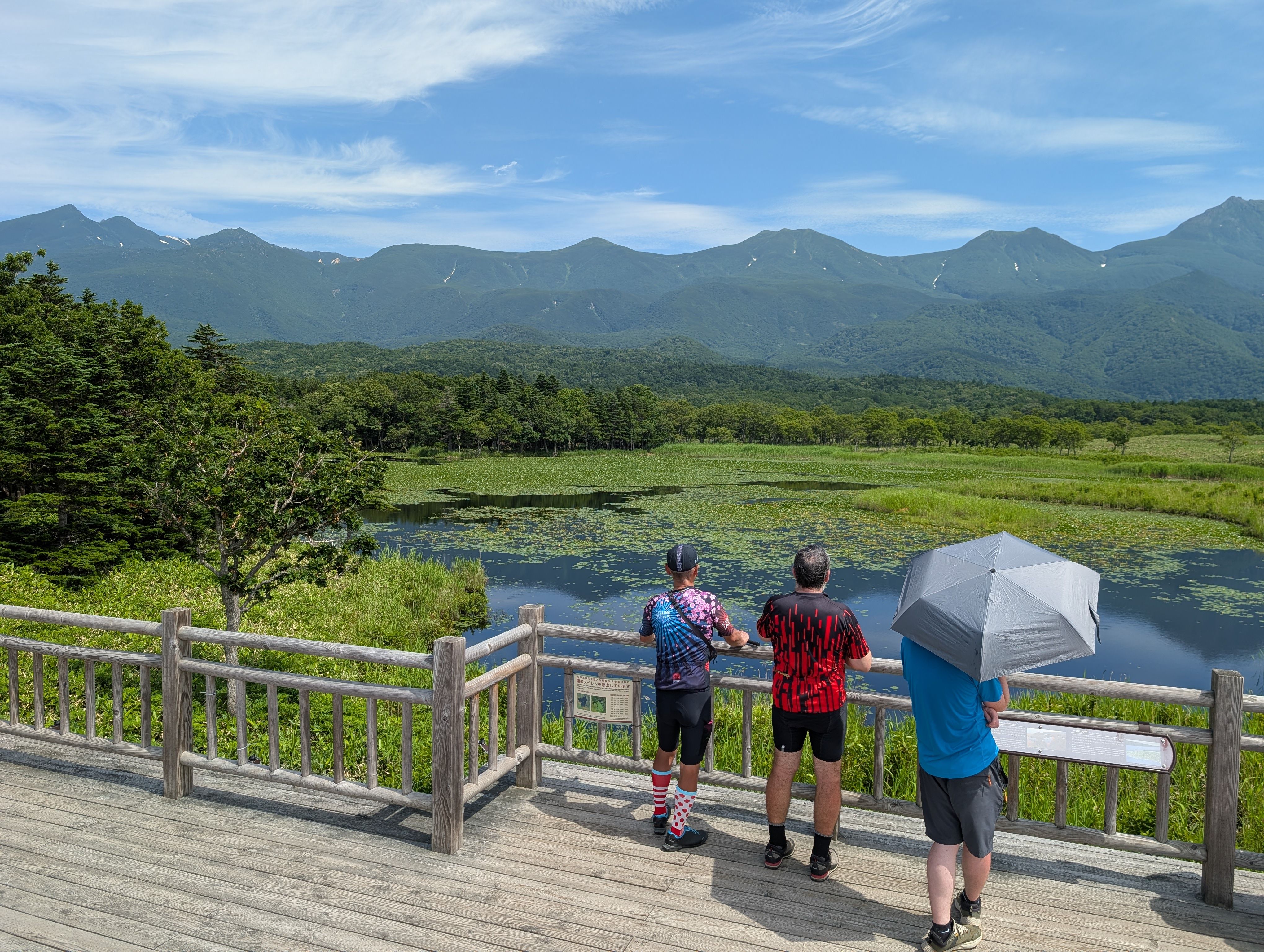 Three people admire the First Lake at Shiretoko Five Lakes. It's a very sunny day and a mountain range is visible on the other side of the lake.