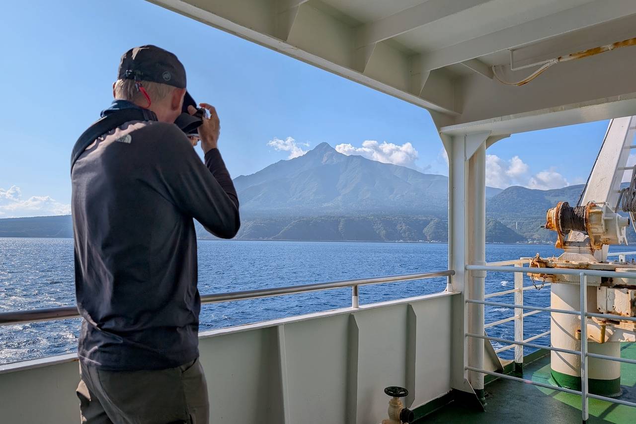 A guest taking a photo from a ferry deck during an Adventure Hokkaido tour, capturing the view of the majestic Mt. Rishiri rising above the deep blue sea under a clear sky.