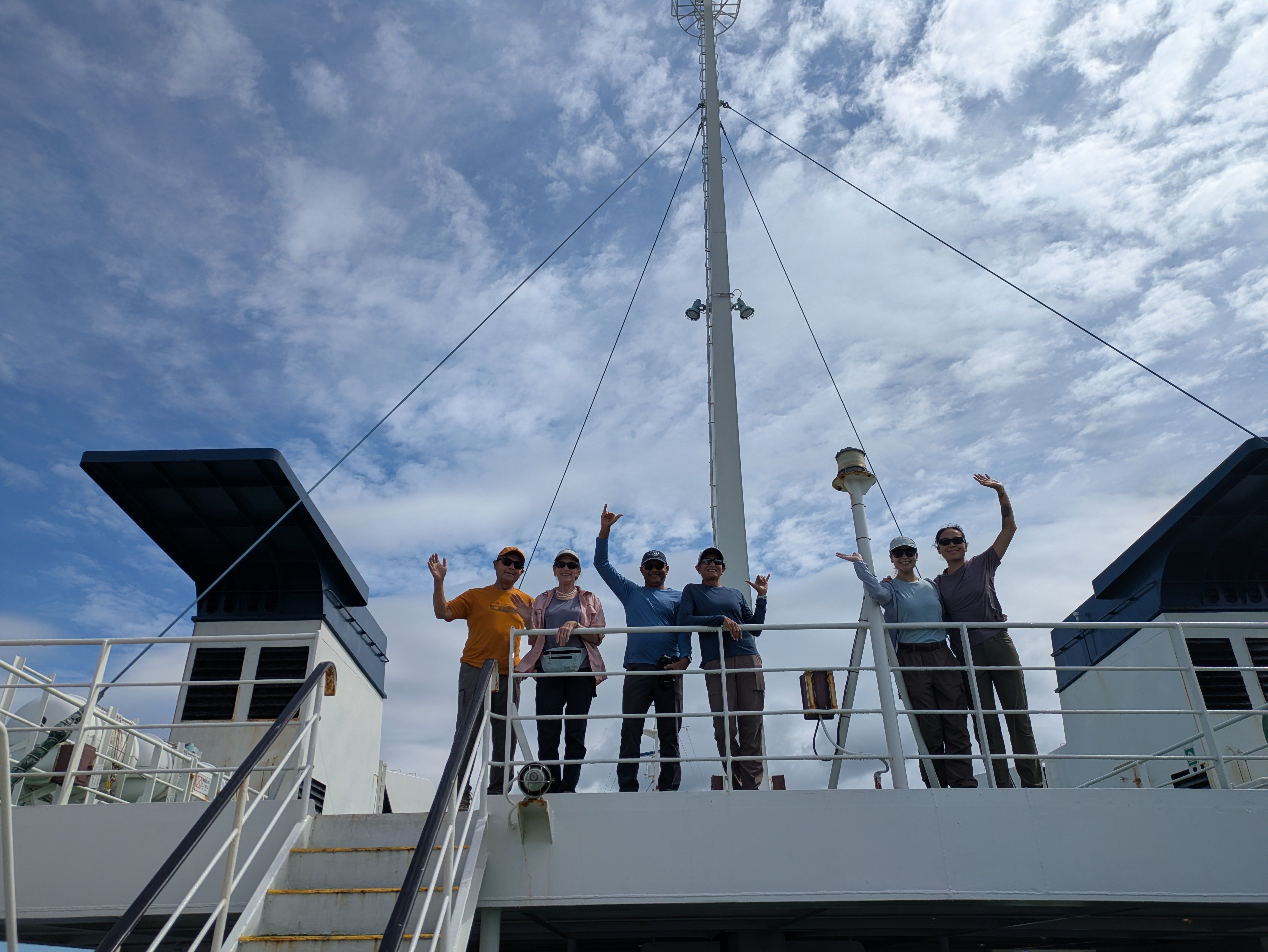 A group of travellers wave at the camera from the upper deck of a ferry. They are framed against scattered wispy white clouds in the sky.