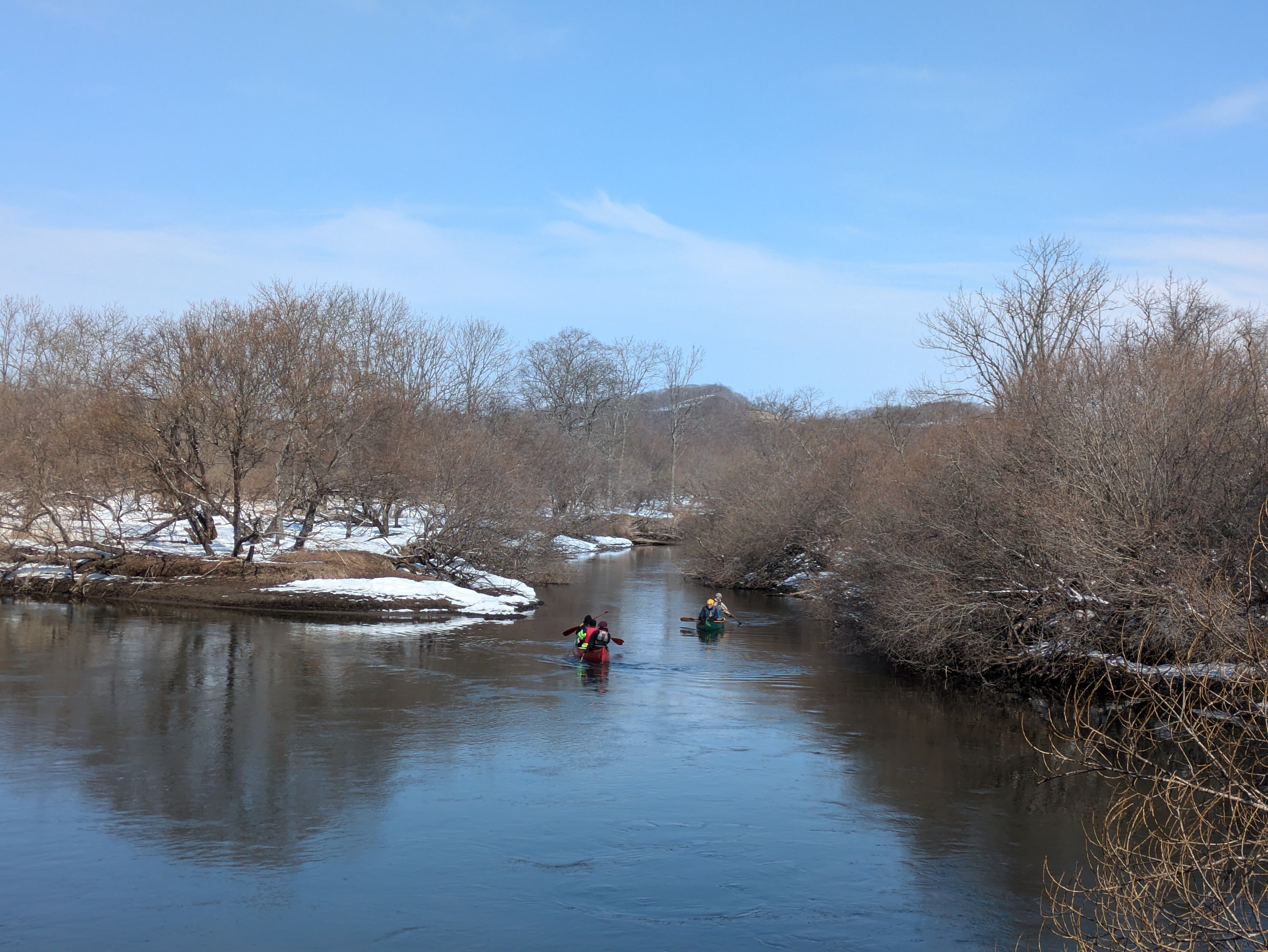 A photo of two canoes on the Kushiro Wetlands, taken from far away. It is a sunny winter day with snow on the riverbank.