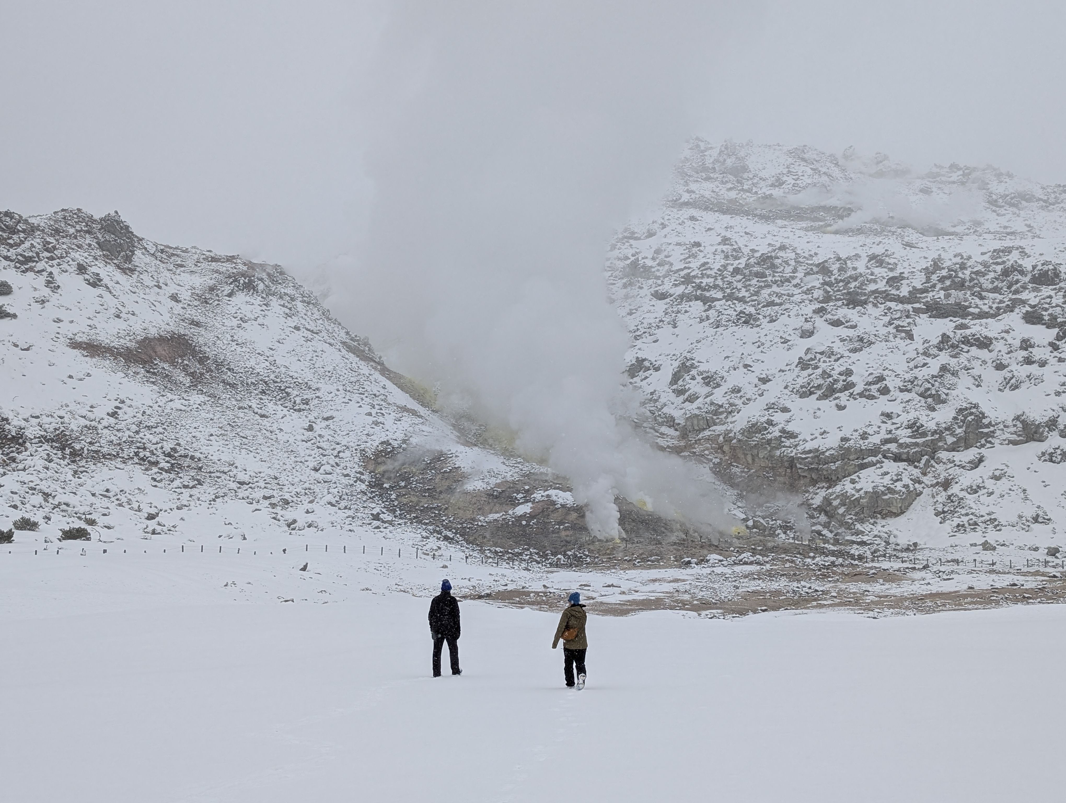 Two figures walk through the snow towards steaming sulphur vents at Mt. Io, Hokkaido, Japan.