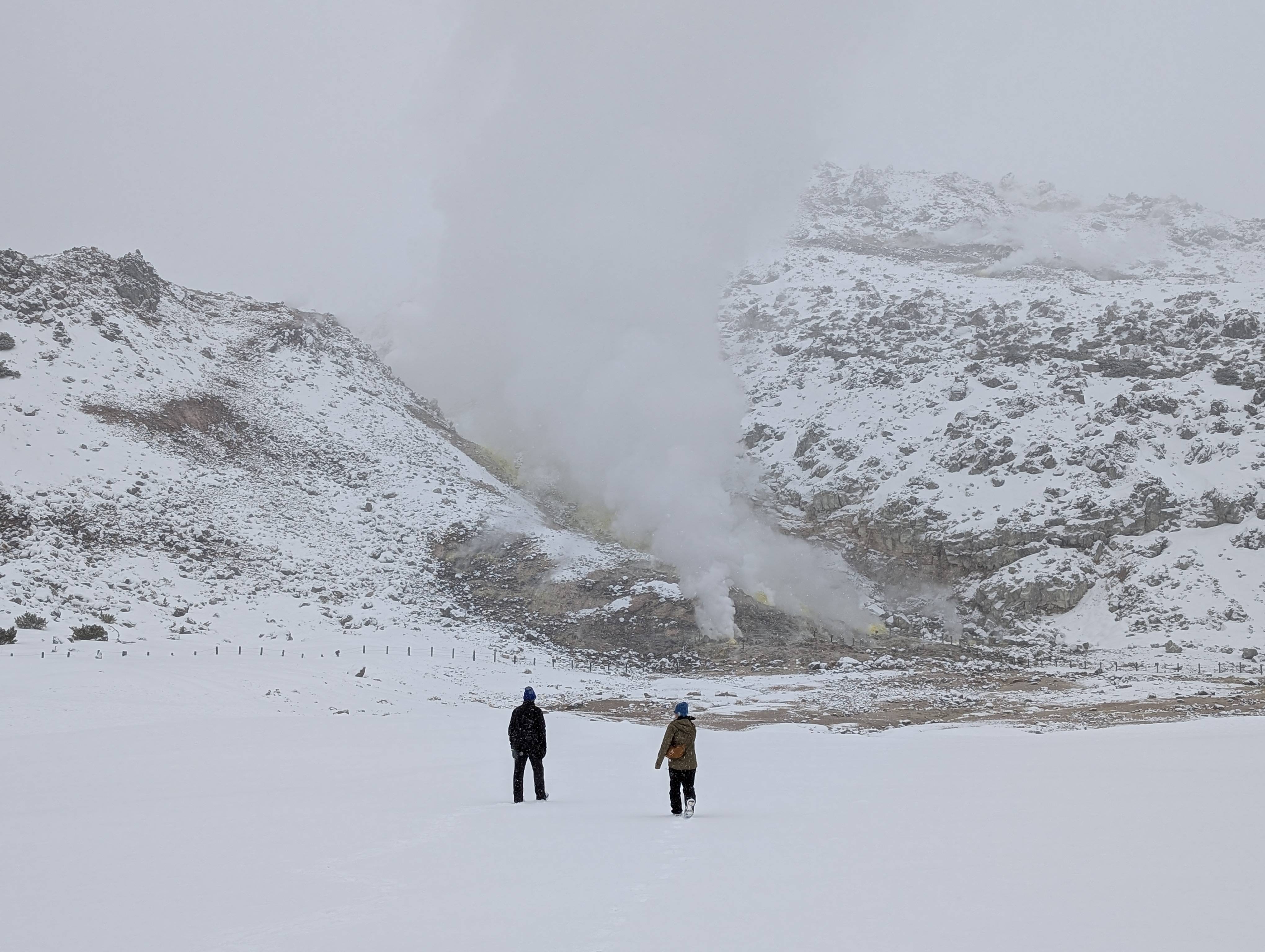 Two figures walk through the snow towards steaming sulphur vents at Mt. Io, Hokkaido, Japan.