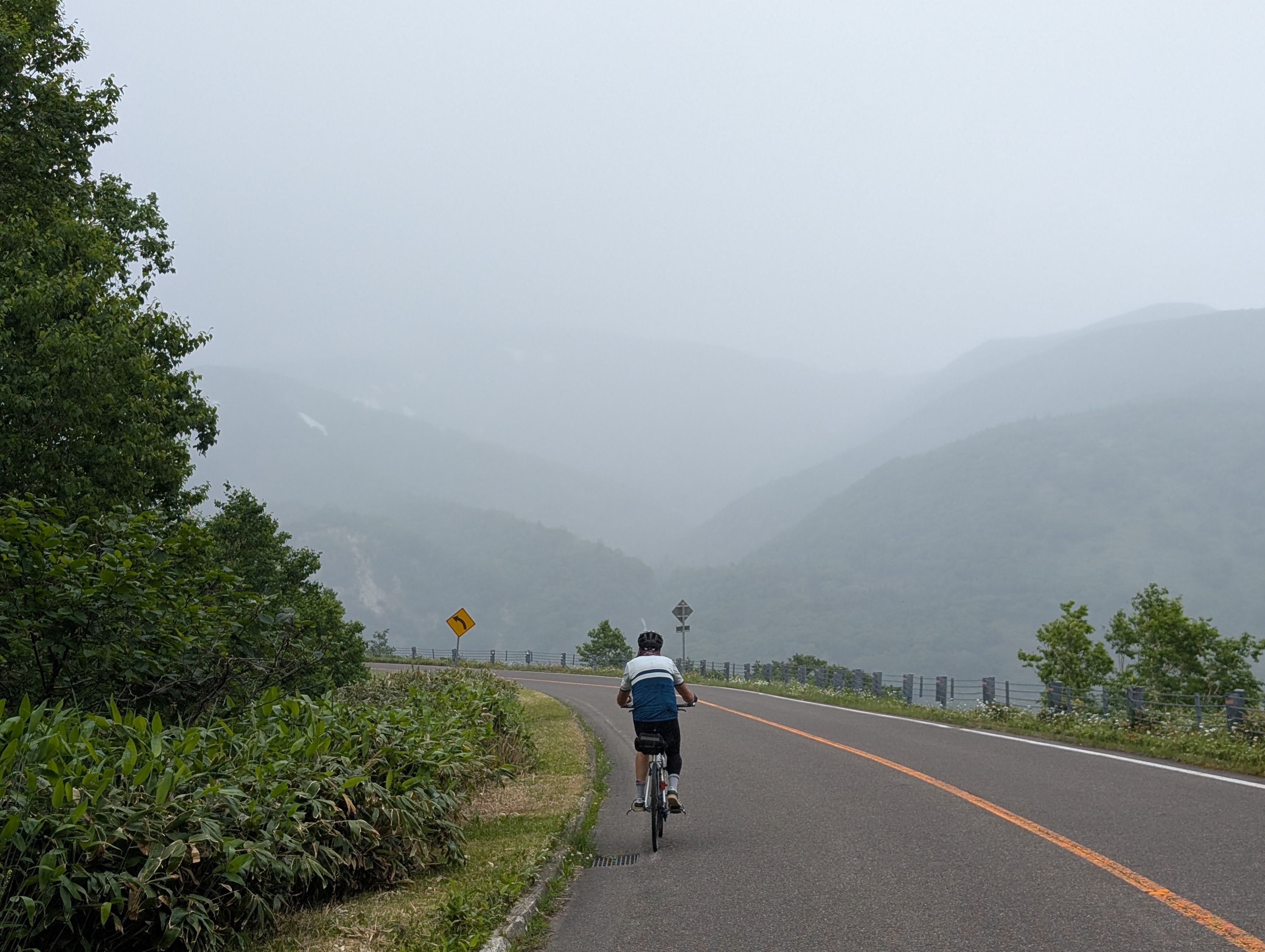 A man on a bike cycles up the Shiretoko Pass. It is very misty and the valley beyond is barely visible.