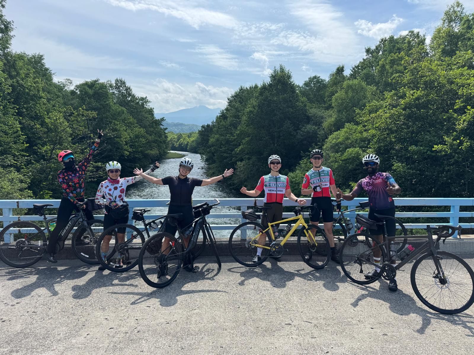 A group of cyclists pose with their bikes on a bridge spanning the Ishikari River in Asahikawa, Hokkaido. It is a sunny day. everyone looks very happy.