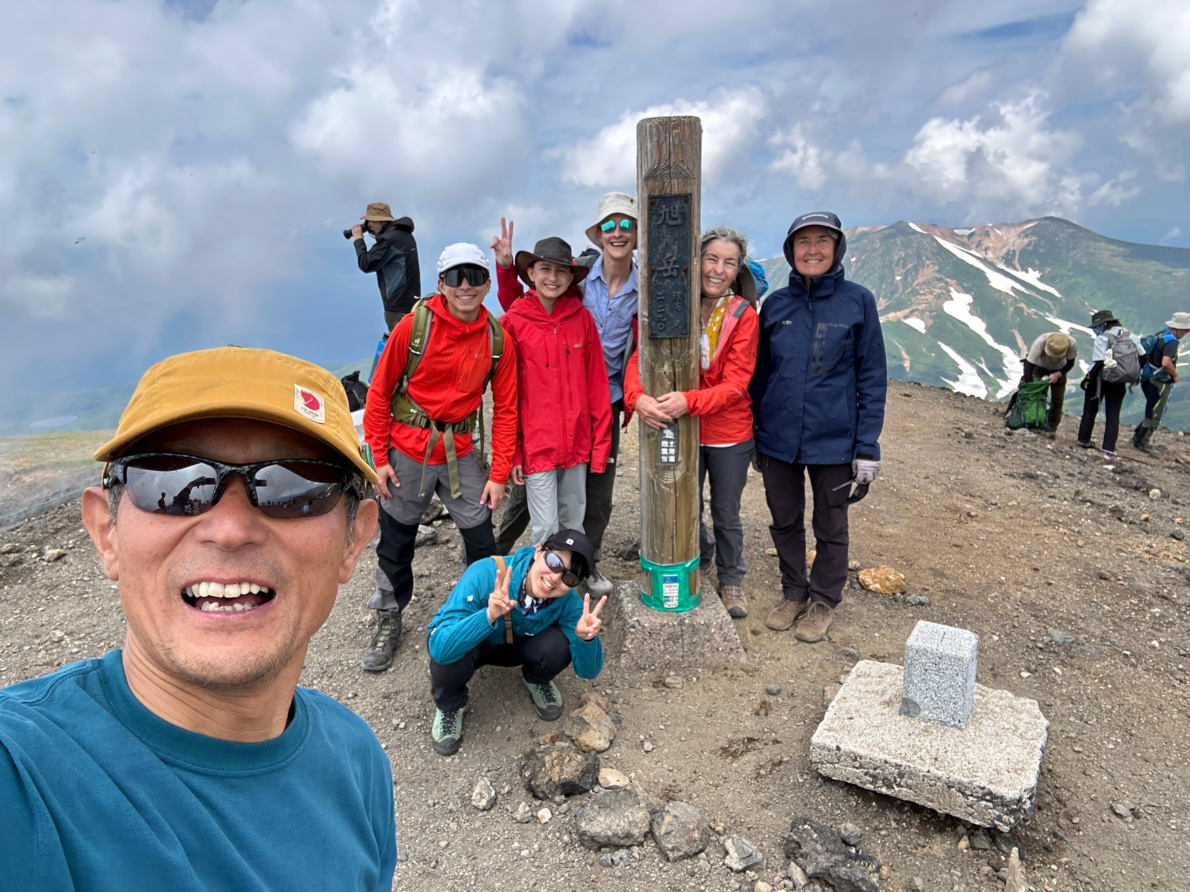 A mountain guide takes a selfie at the summit of Mt. Asahidake with his guests and co-guide behind him. A pillar in the ground reads "Mt. Asahidake: 2,290m" in Japanese.