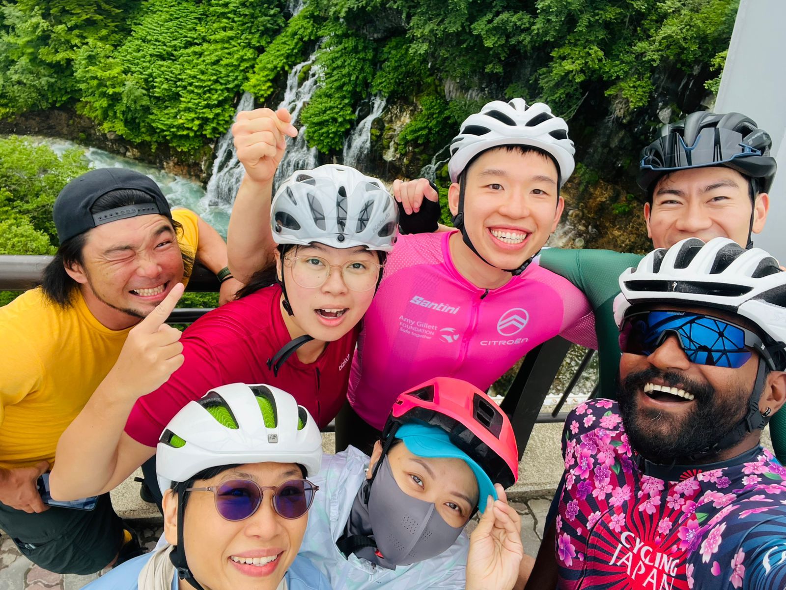 A group of seven cyclists take a selfie in front of Shirahige Waterfall. They are all smiling and looking excited. They wear cycling gear and helmets.