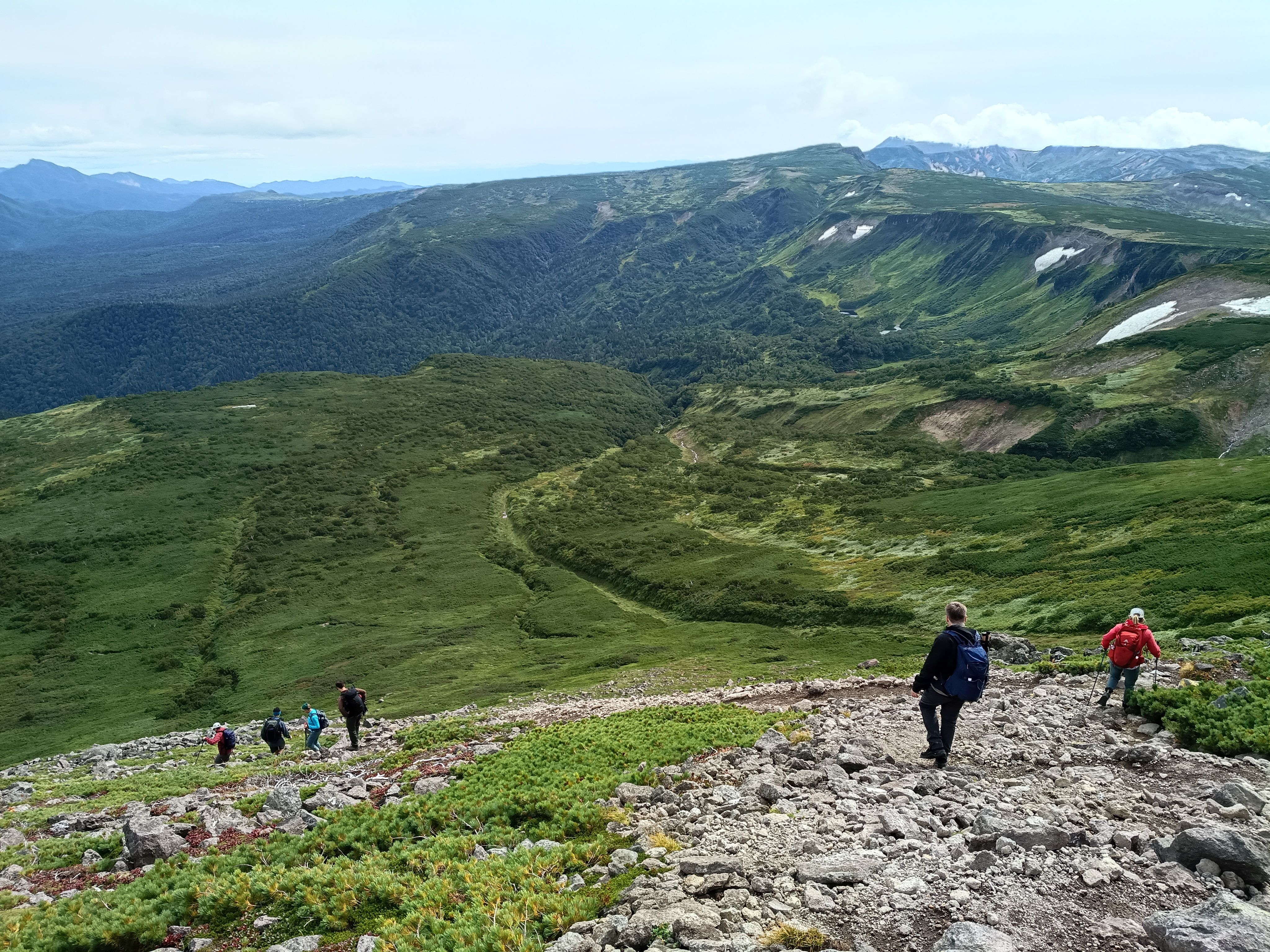A group of hikers on a tour descend a rocky trail on Mt. Midori in Daisetsuzan National Park, overlooking a vast, green mountain valley.