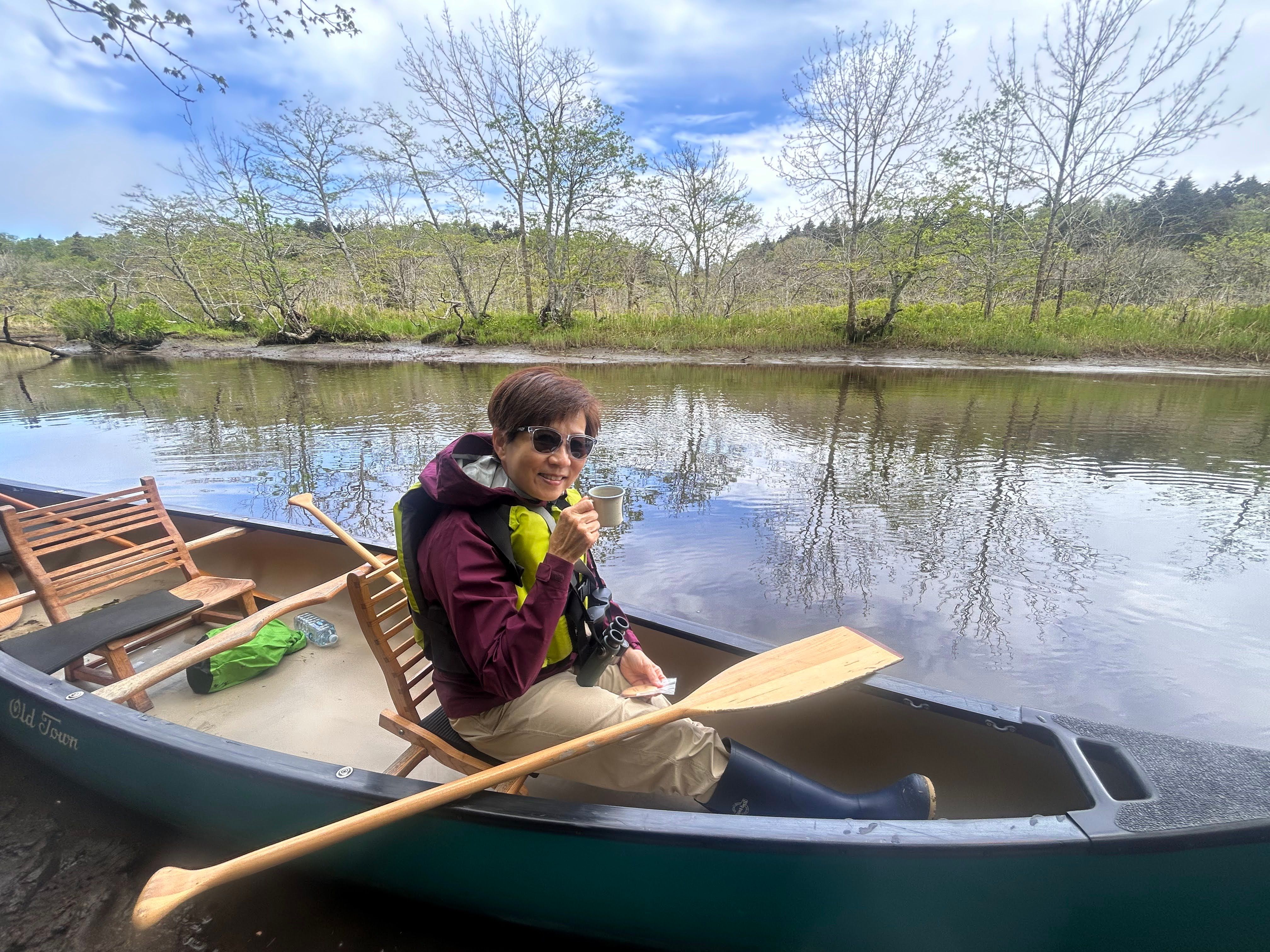 A woman sitting in a canoe and wearing sunglasses raises a coffee cup to the camera and smiles. It is a sunny day.