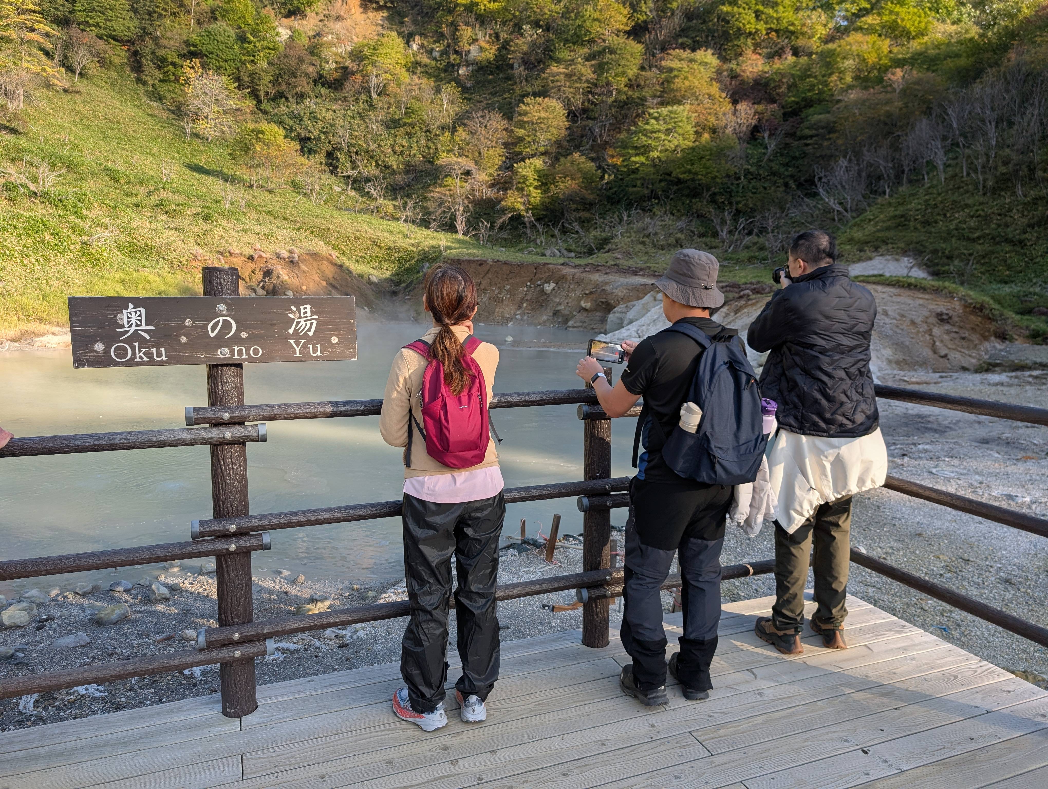 Three people stand at a fence next to Oku no You hot spring at Noboribetsu Onsen, Hokkaido. Beyond the fence is a hot spring pond. It is a milky blue-grey colour.