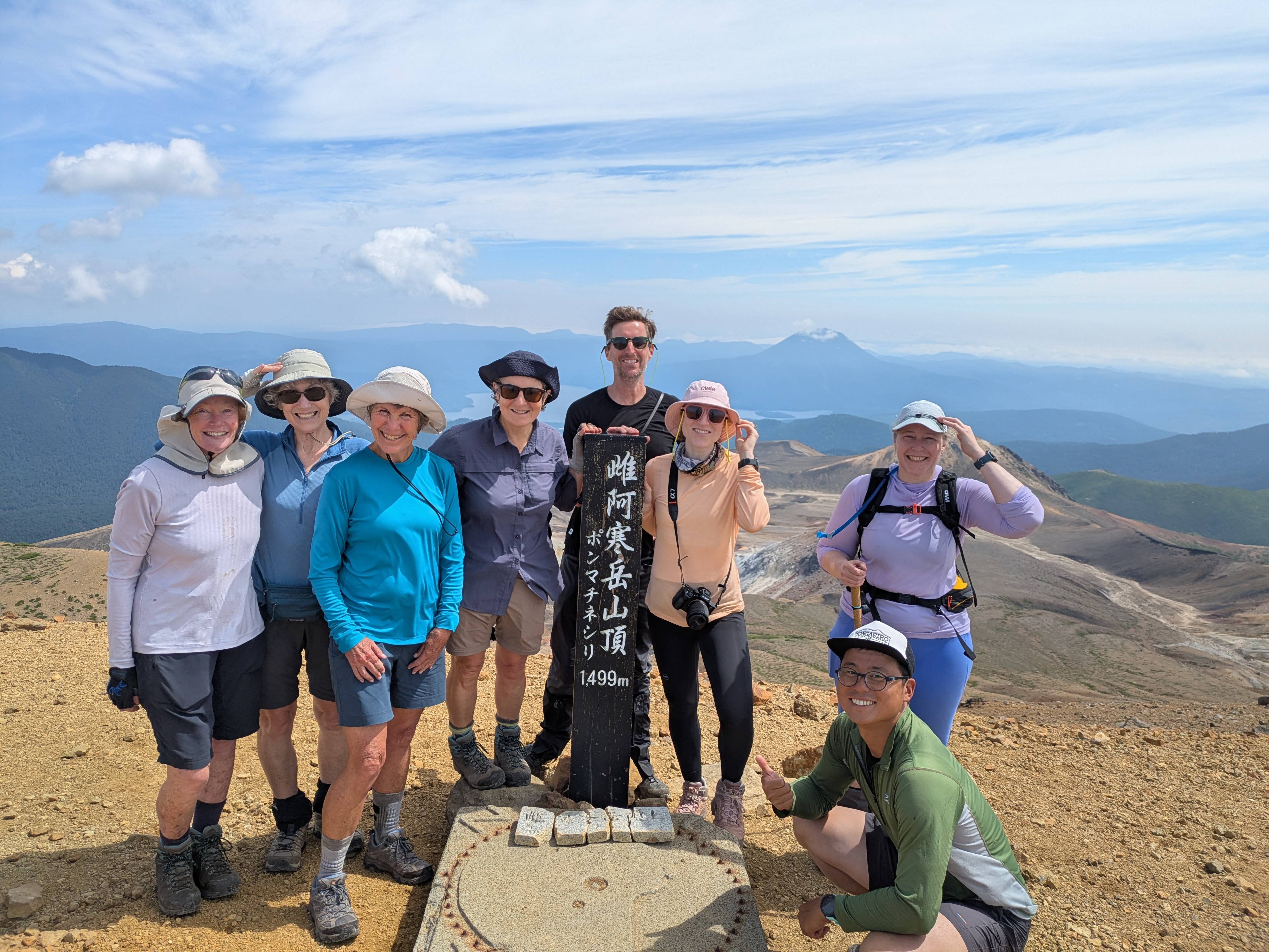 A group of hikers smile at the summit of Mt. Meakan in Hokkaido. They are standing around a pole which reads in Japanese, "Mt. Meakan Summit - 1499m".