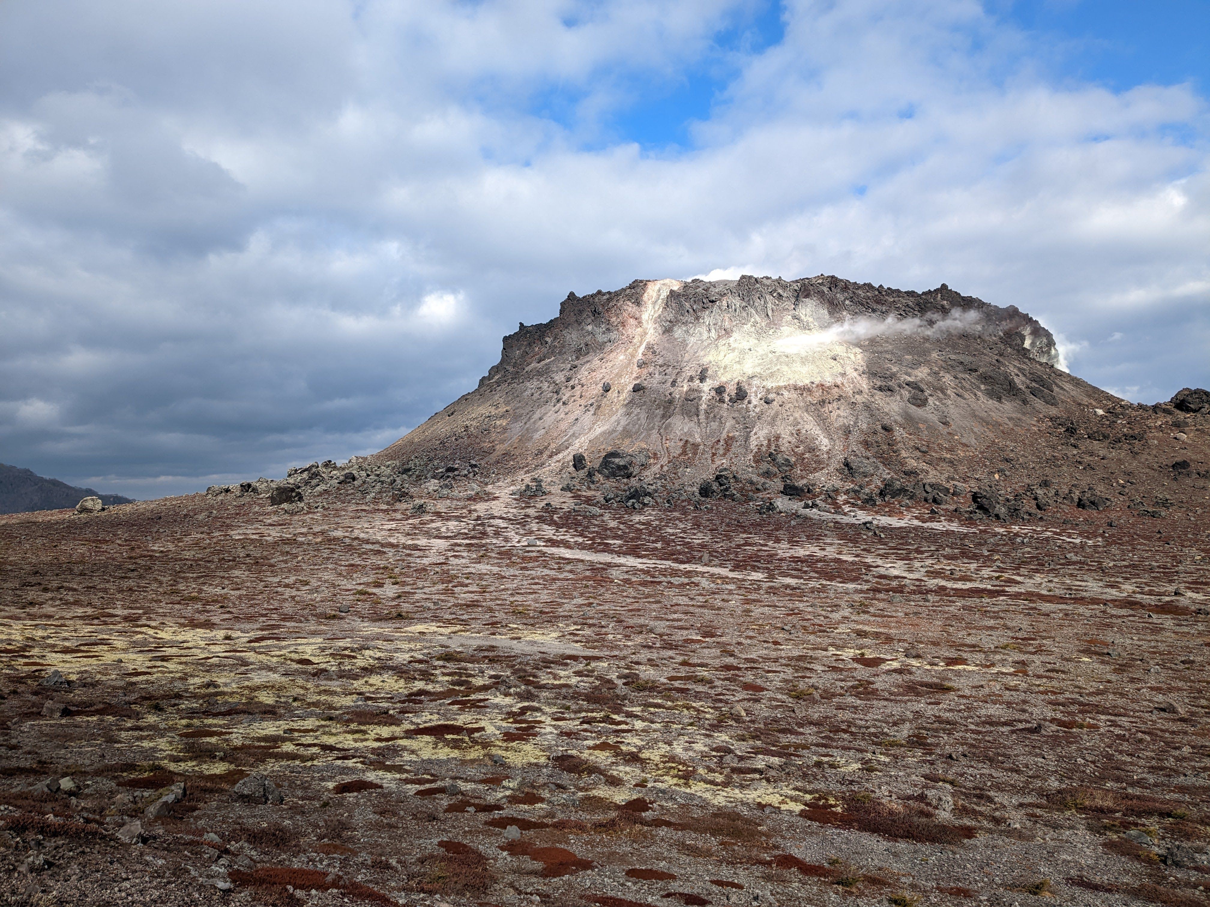 The rugged, steaming summit of Mt. Tarumae in Shikotsu–Toya National Park rises above a desolate plain of rock and patches of low plants. The plants have turned brown indicating it is late in the year. Plumes of gas vent from brilliant yellow and white sulphur deposits that stain the dark, craggy volcanic dome.