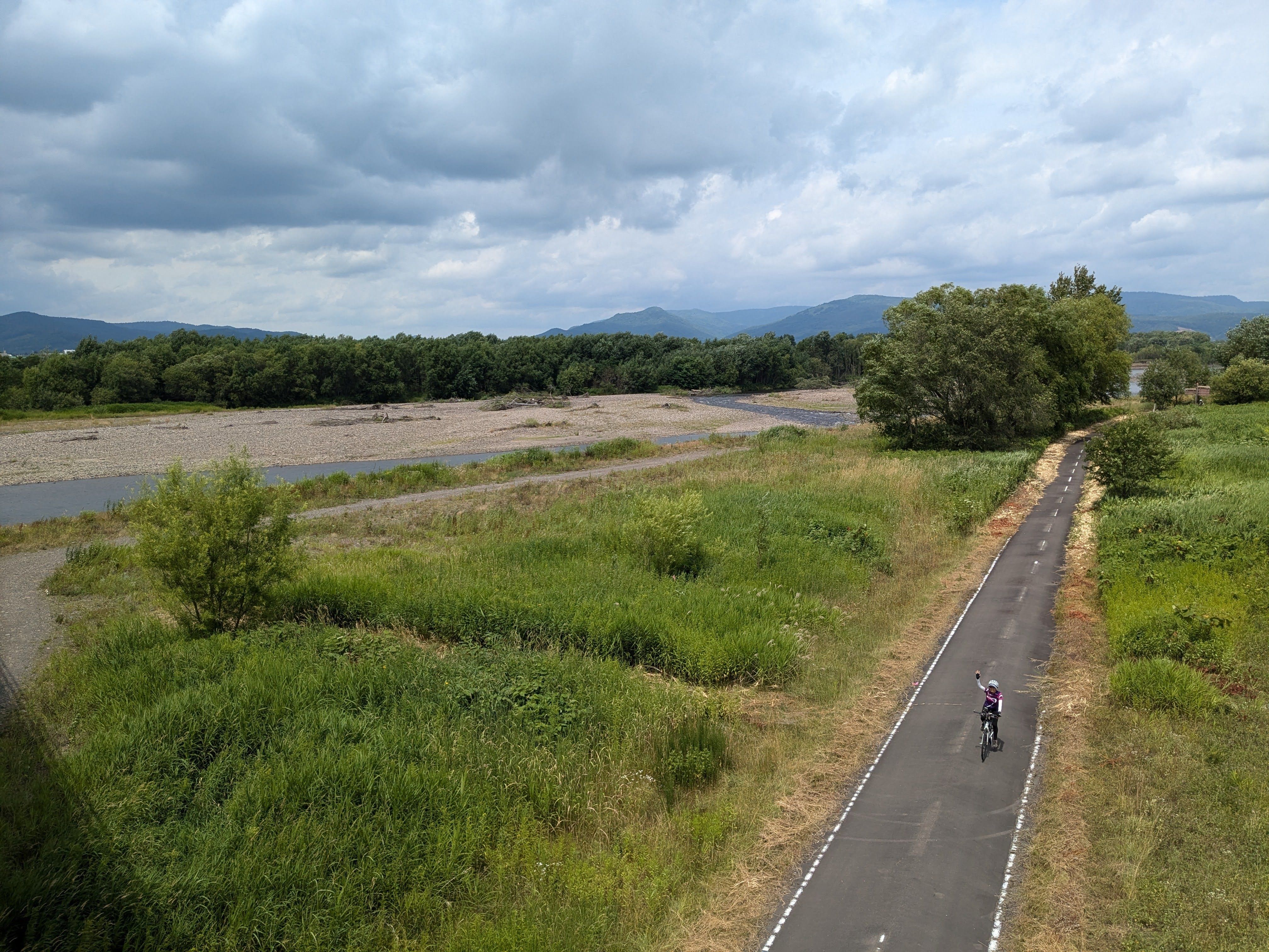 An aerial view of a cyclist on a cycling path alongside a river in Asahikawa, Hokkaido. He is punching the air in victory. It is a cloudy day.