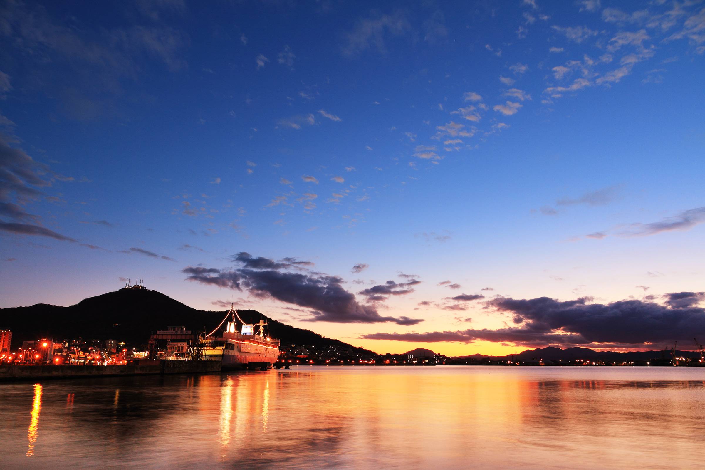 A nighttime view of Hakodate Bay. The sun is setting and reflecting off the water, along with the lights of ships and buildings. Mt. Hakodate is visible in the background.
