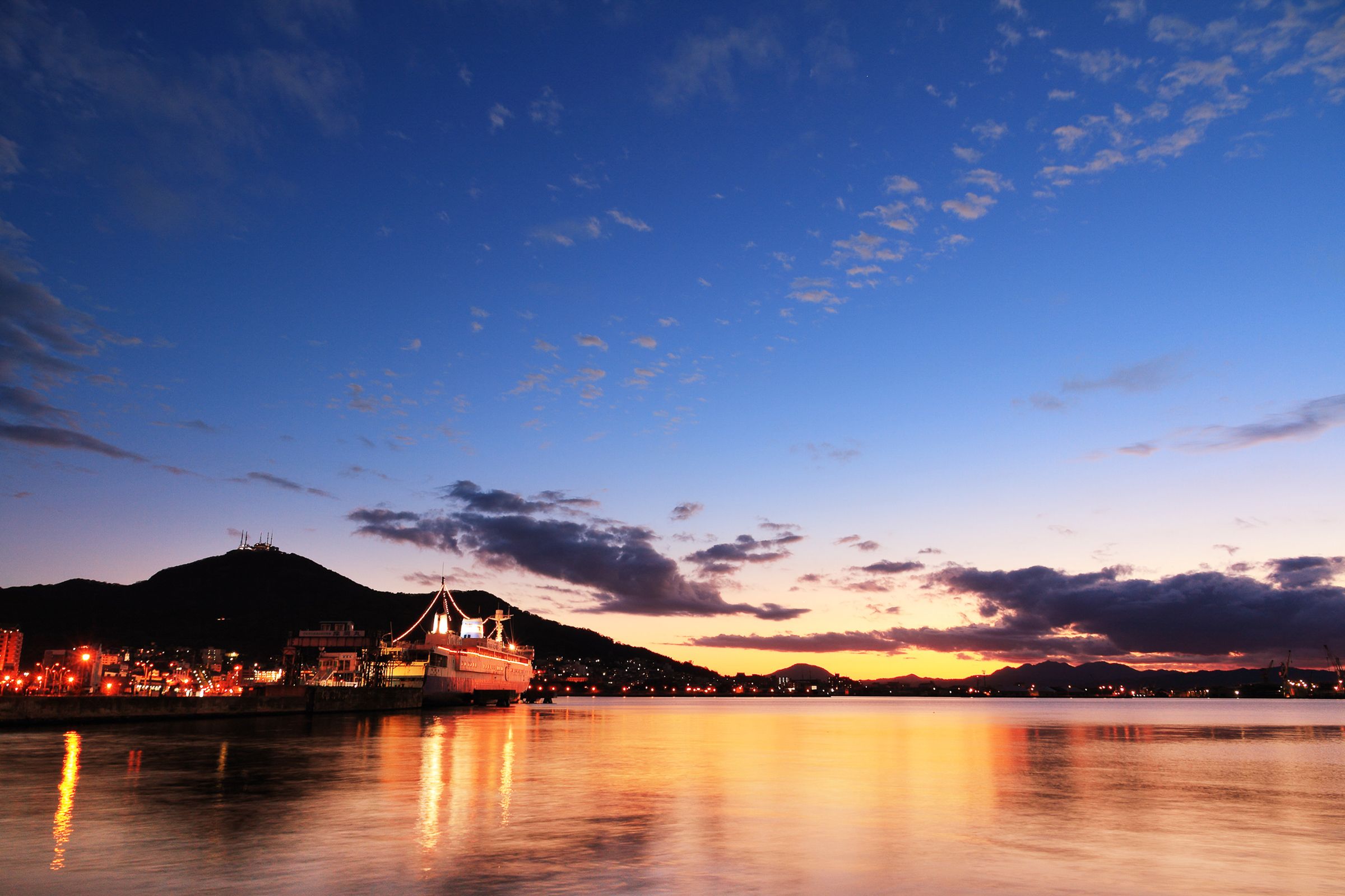 A nighttime view of Hakodate Bay. The sun is setting and reflecting off the water, along with the lights of ships and buildings. Mt. Hakodate is visible in the background.