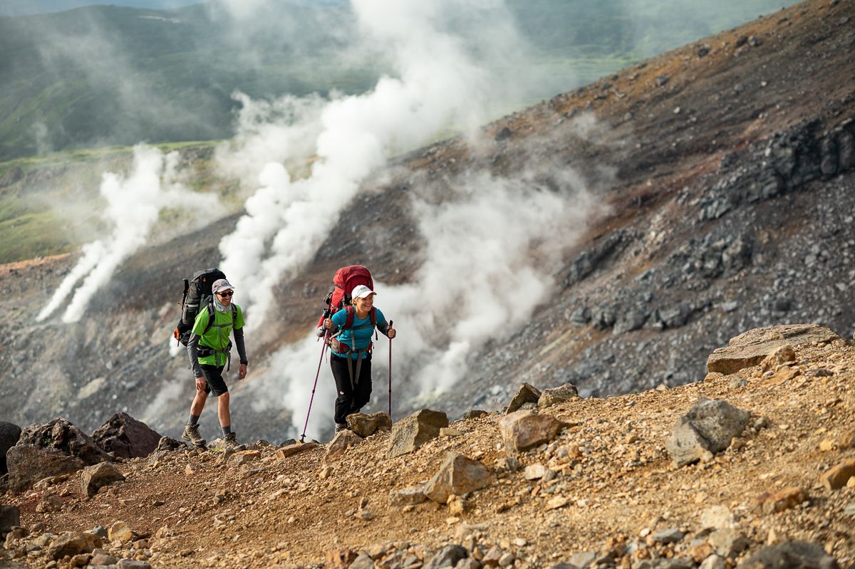 Two hikers with large packs ascend the steep, rocky ridge of Mt. Asahidake in Daisetsuzan National Park. The volcanic landscape surrounds them, with powerful plumes of white steam billowing from fumaroles on the mountainside, creating a hazy and primordial atmosphere under a bright sky.