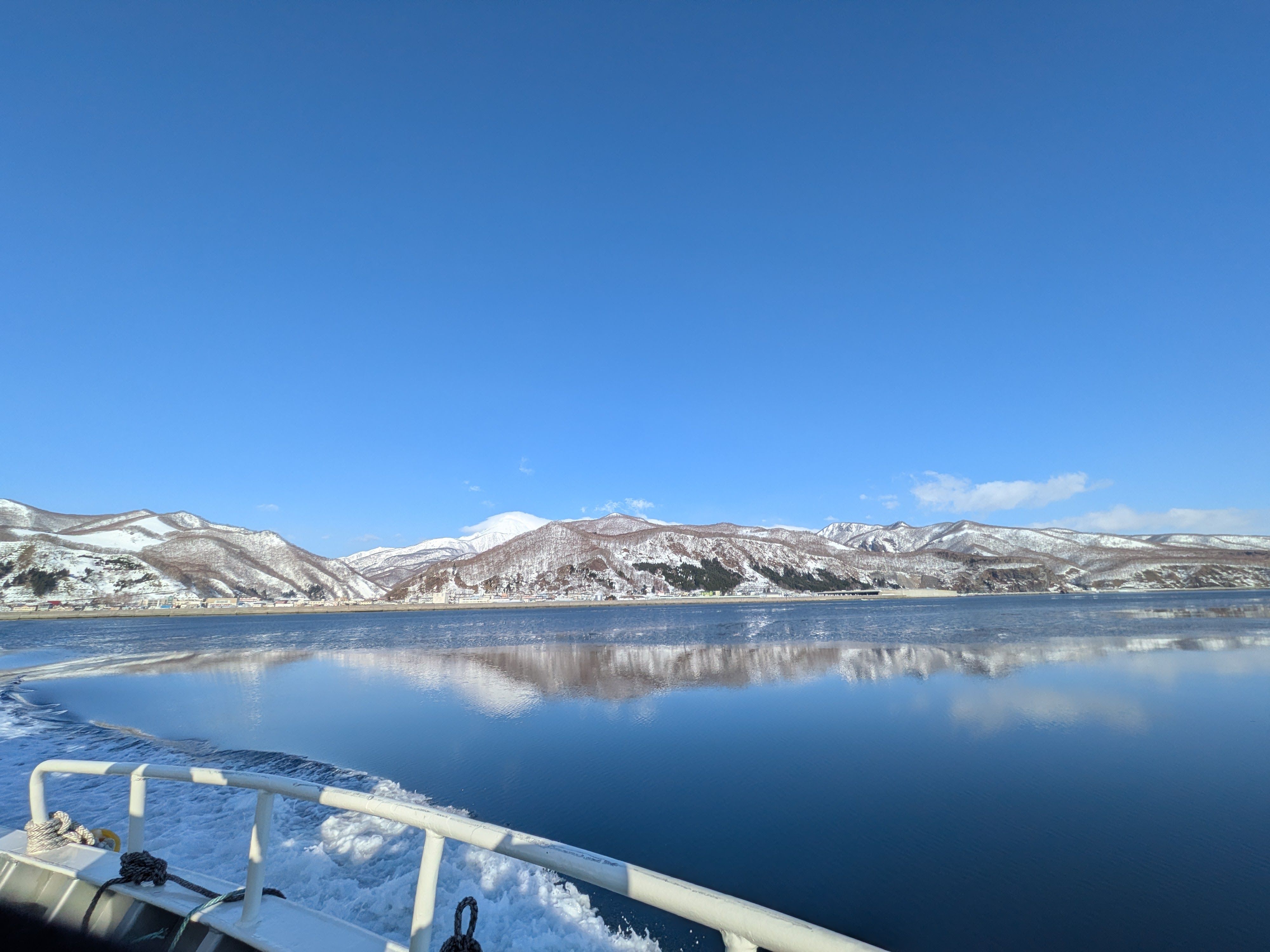A photograph of the ocean from the deck of a nature cruise ship. It is a very still day and the water's surface looks like a mirror, perfectly reflecting the mountains and the blue sky.