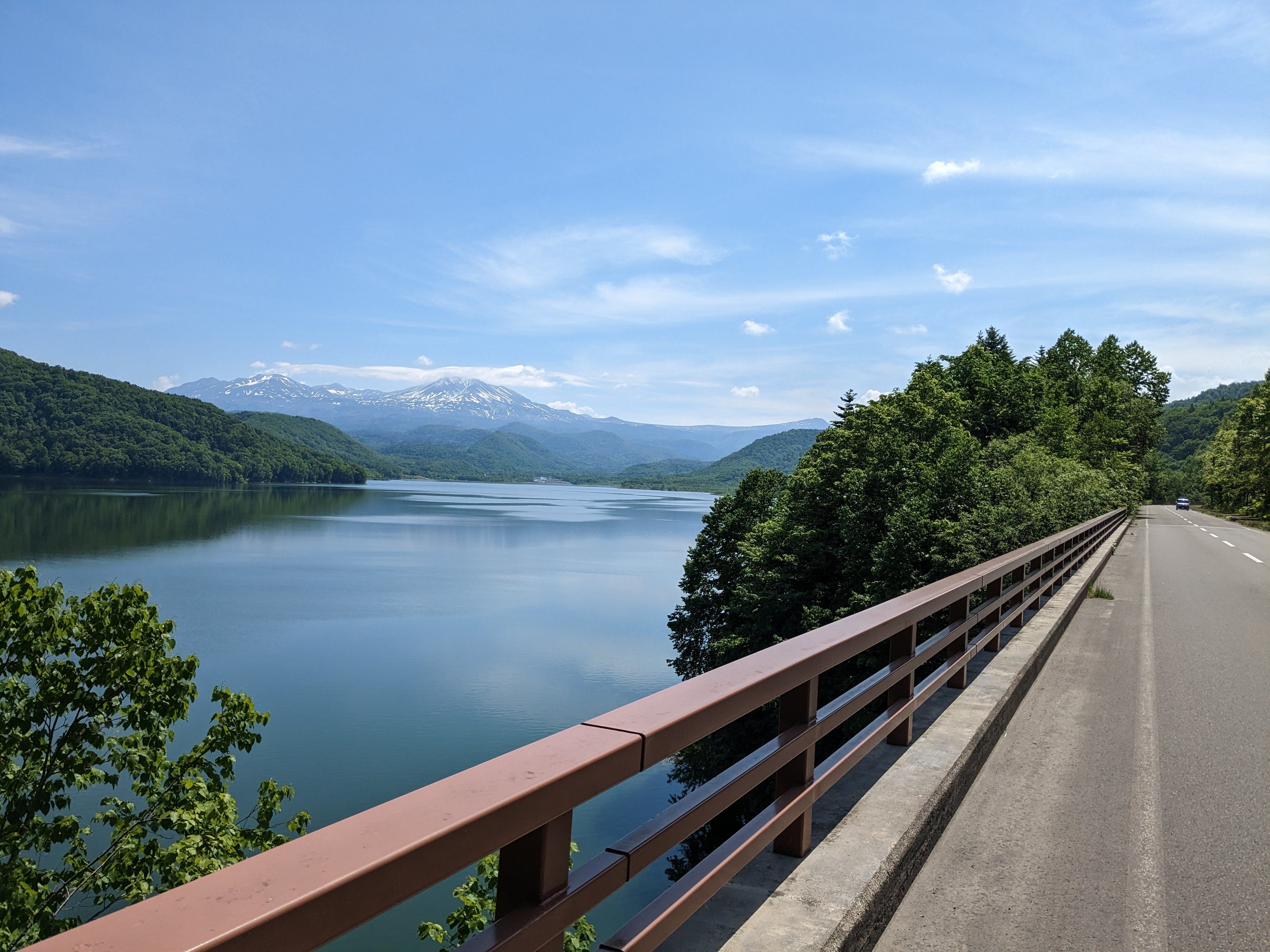 A view of Mt. Asahidake, the tallest mountain in Japan, from a bridge over the far side of Chubetsu Lake. It is a very sunny day and the mountain is clearly visible beyond the far lake shore.
