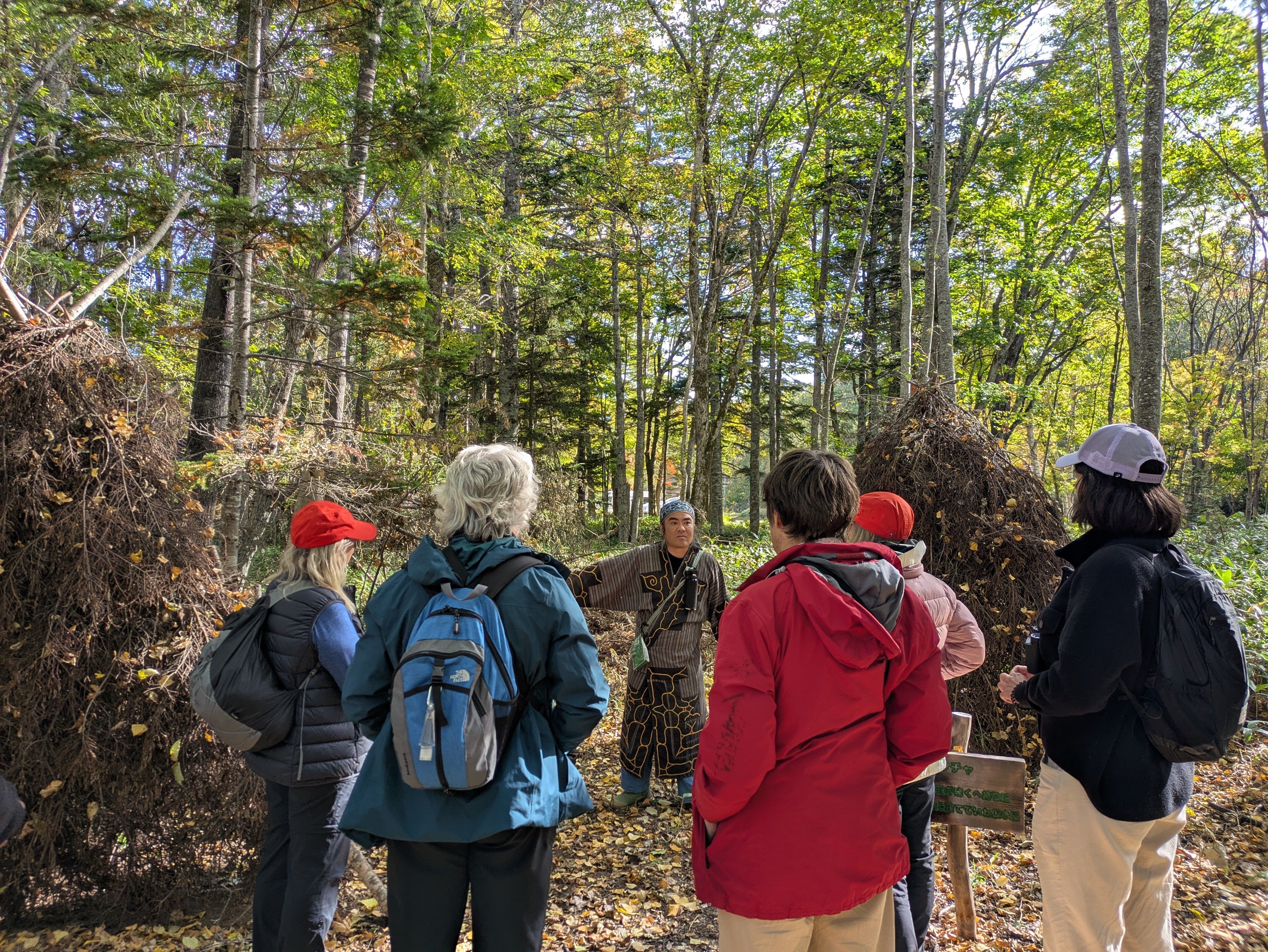 A man dressed in the traditional garb of the Ainu, Hokkaido's indigenous people, addresses a crowd of visitors. There are shelters made of piled up tree branches around him.