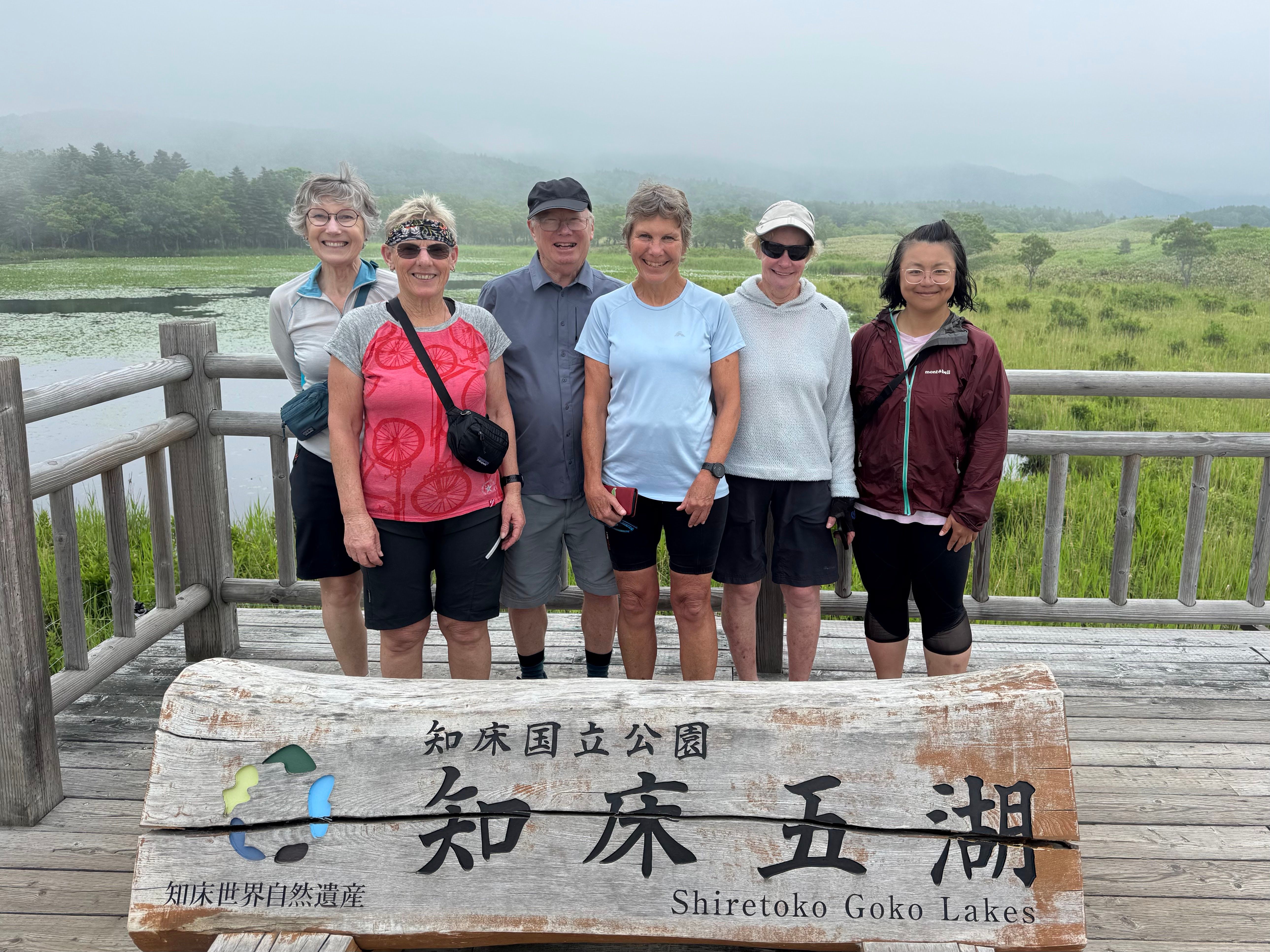 A group of people stand behind a sign and smile at the camera. The sign reads, in Japanese, "Shiretoko National Park - Shiretoko Five Lakes". Lake number five is visible behind them.