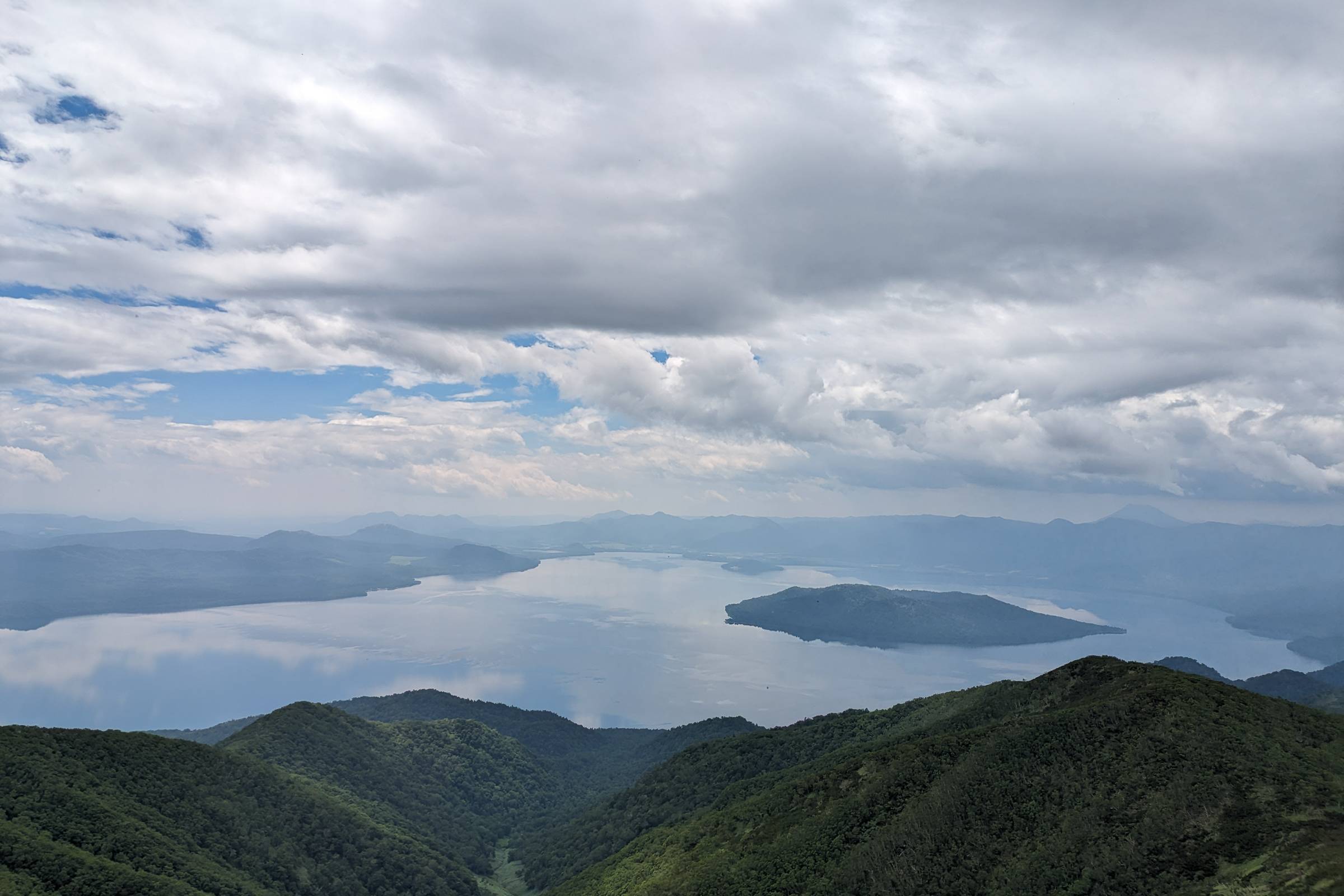 The view of Lake Kussharo from Mt. Mokoto. It is a partially cloudy day and an island is visible in the middle of the lake.