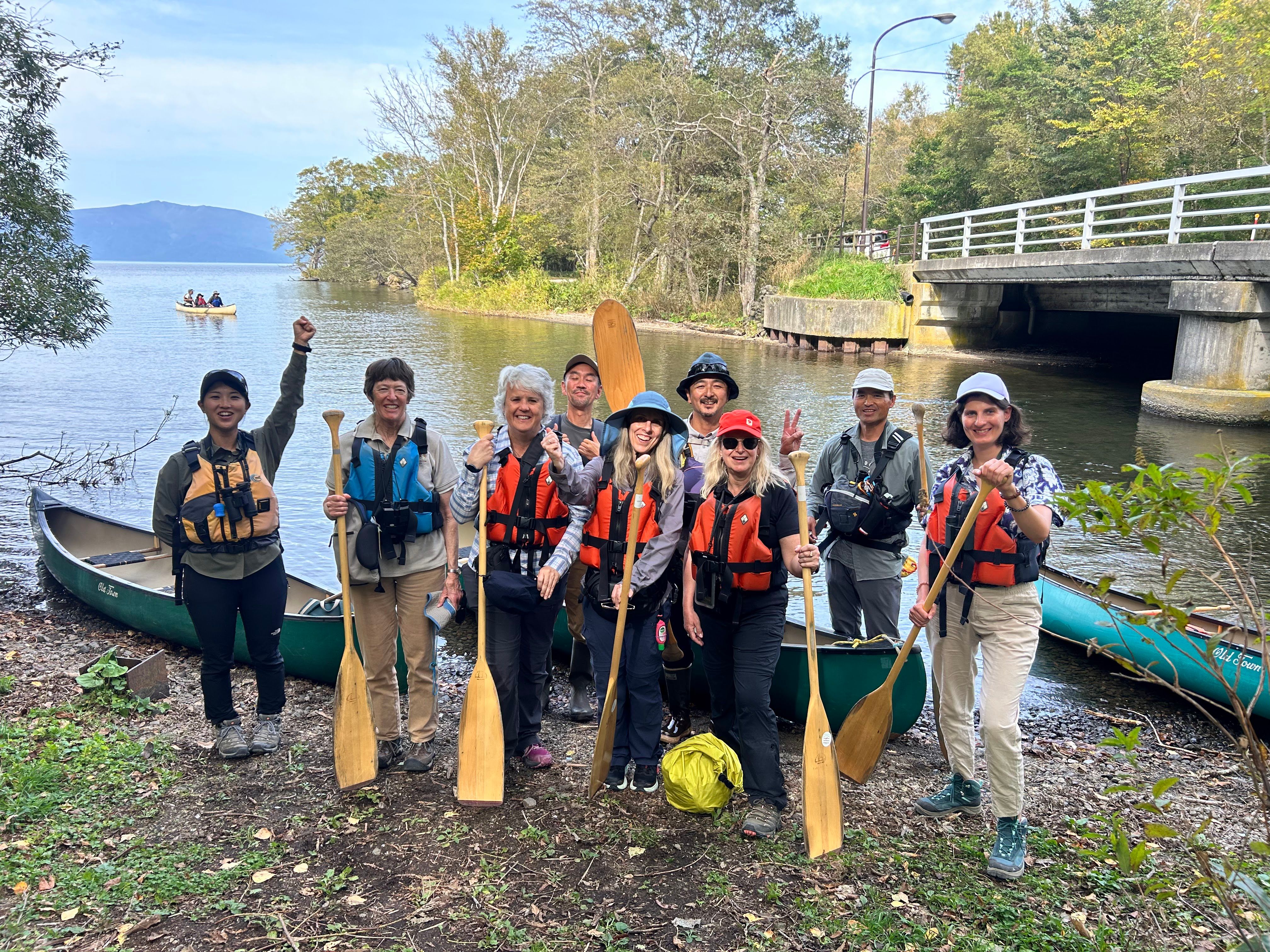 A group of guests and guides pose with oars in front of canoes on the shore of Lake Kussharo. They are all giving thumbs-up, peace signs, punching the air or otherwise looking enthusiastic or excited.
