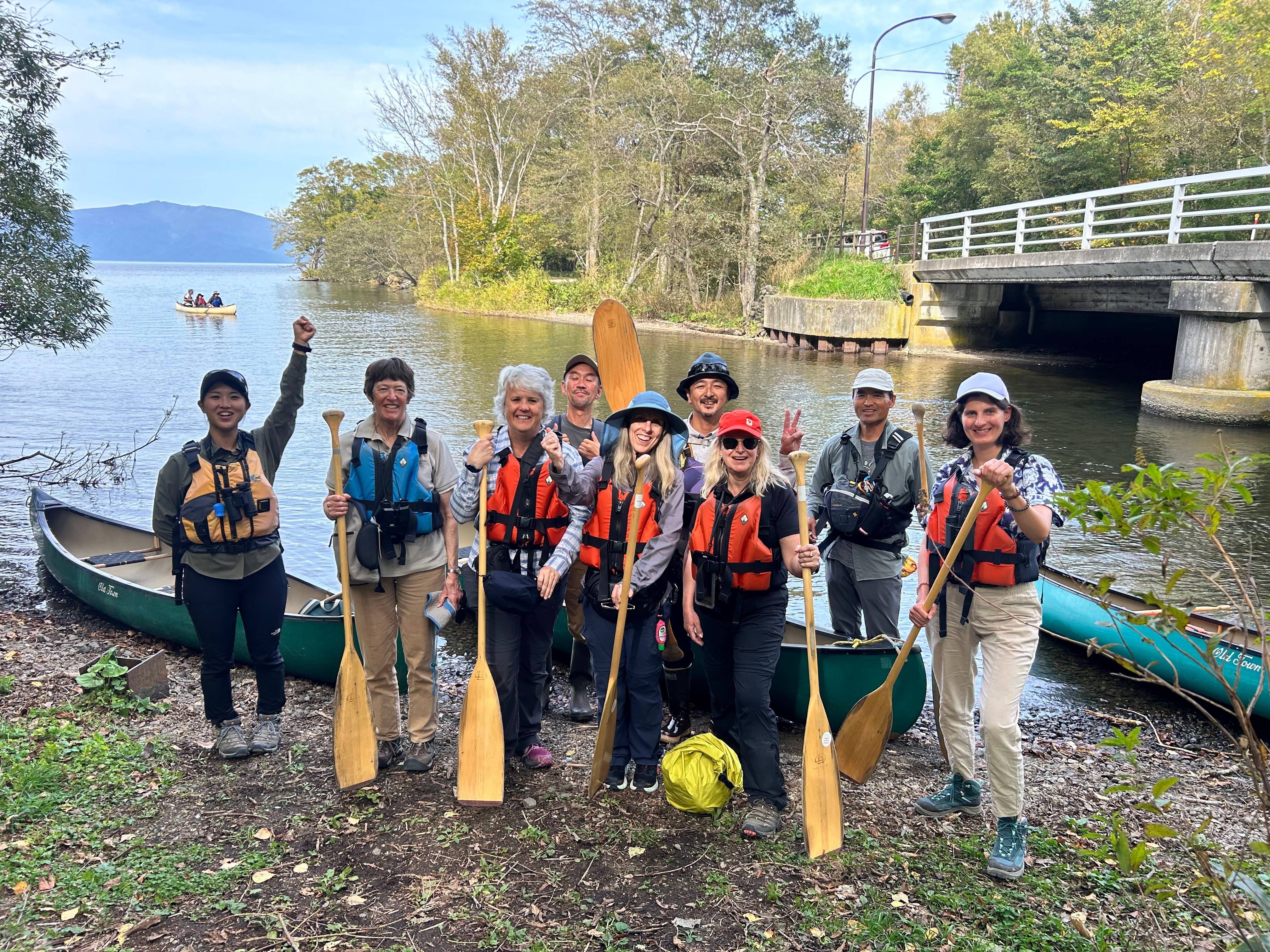 A group of guests and guides pose with oars in front of canoes on the shore of Lake Kussharo. They are all giving thumbs-up, peace signs, punching the air or otherwise looking enthusiastic or excited.