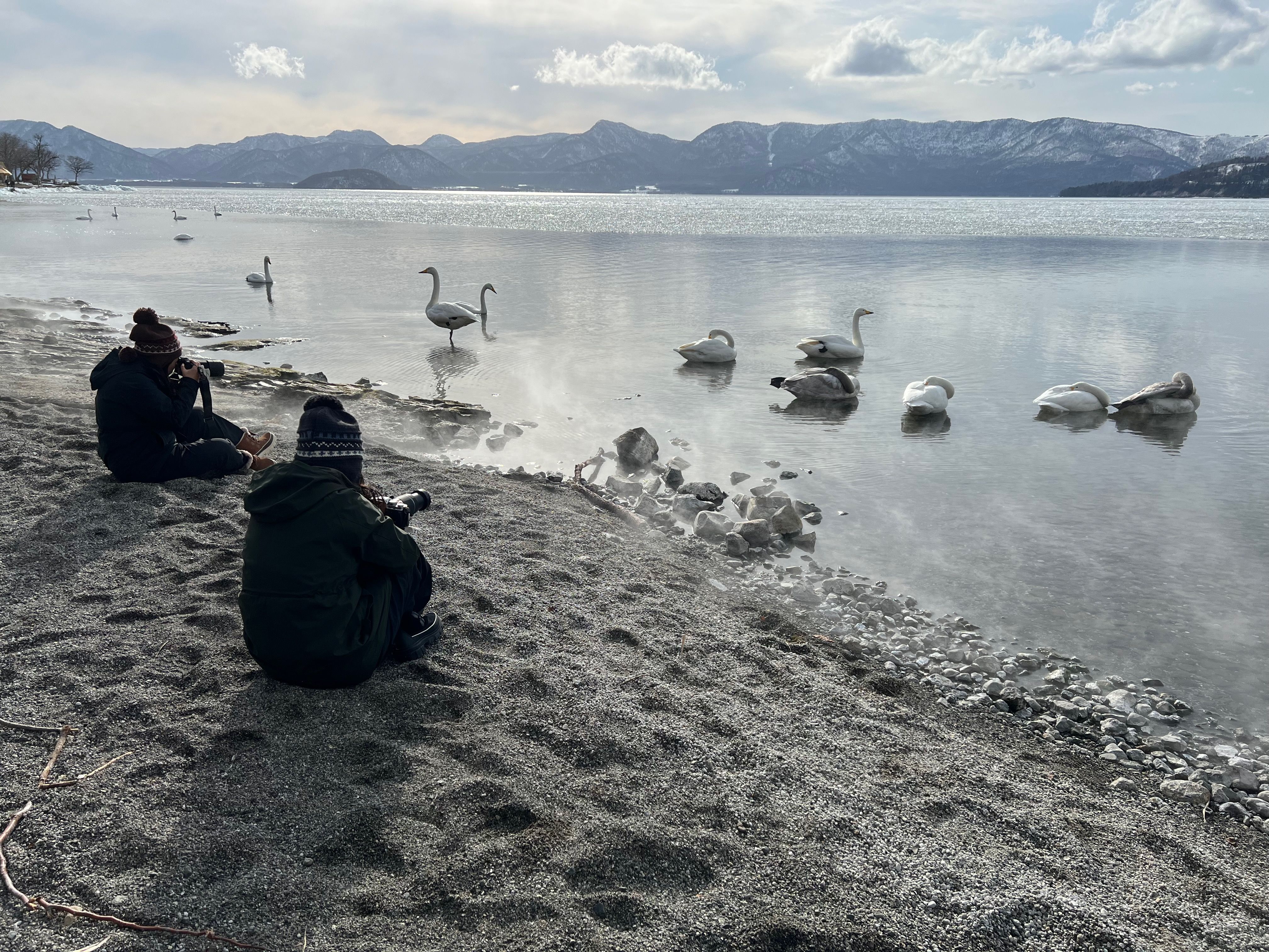Two women with cameras sit on the shores of Lake Kussharo, Hokkaido in the afternoon. There are a group of swans in the waters in front of them. The shore of the lake sits above a hot spring, so steam rises from the water's surface.