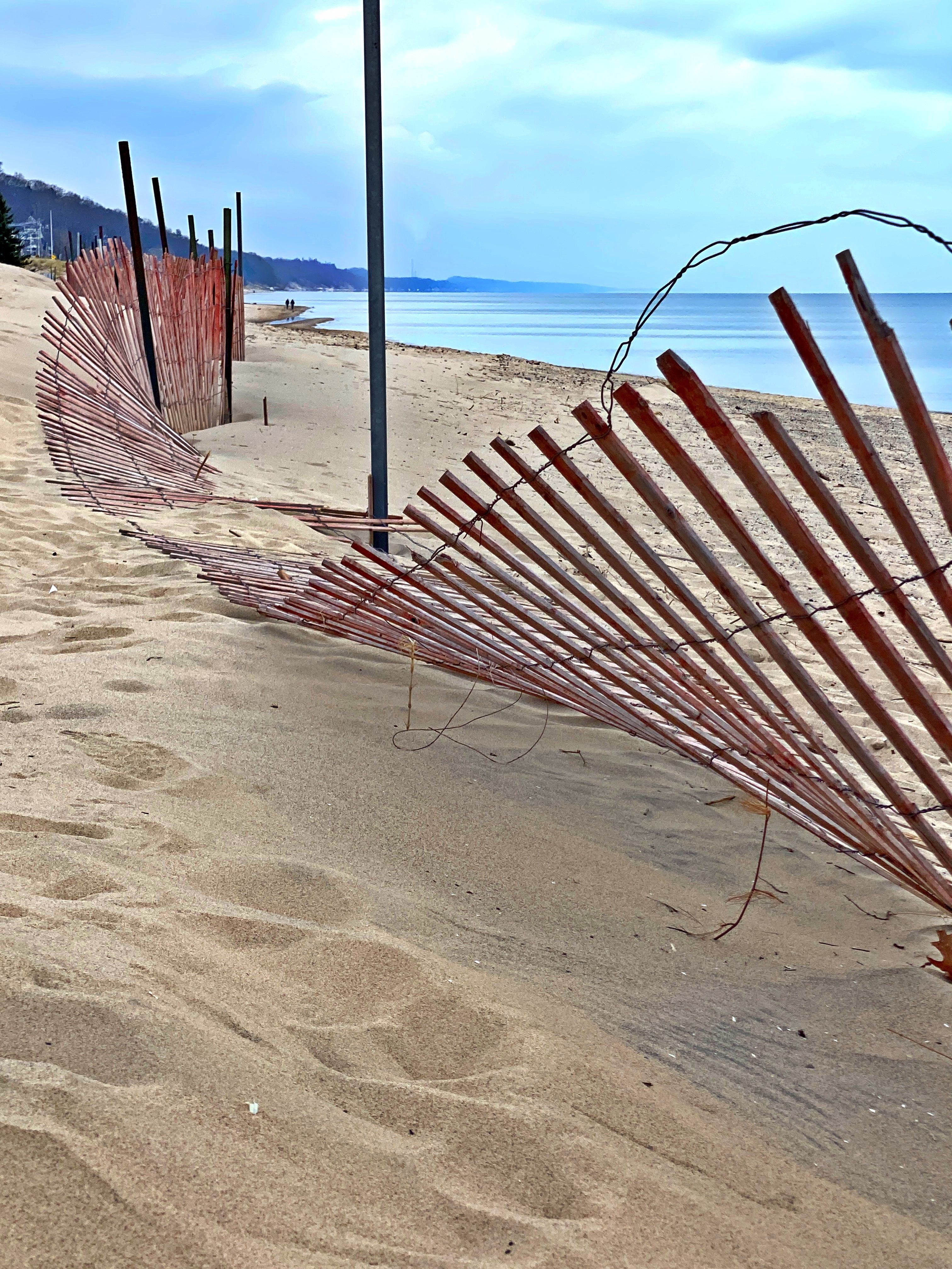 A weathered wood dune fence curves along a sandy beach beneath a pale blue sky
