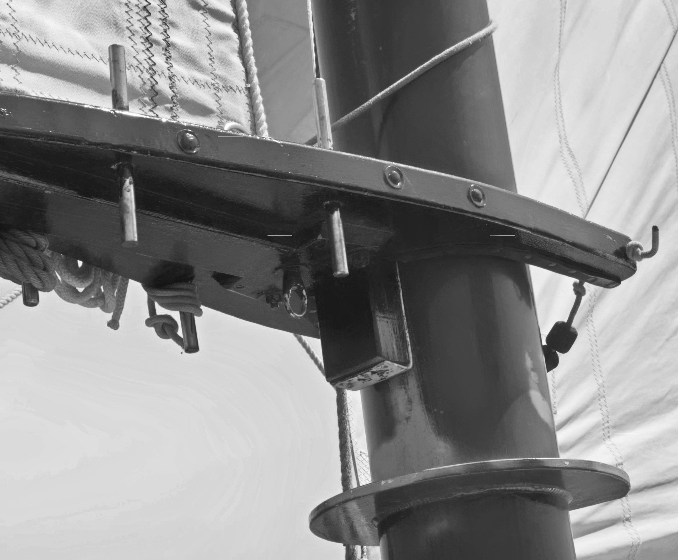 Black-and-white close-up of a boom fitting and belaying pins where mast meets spar, sail behind