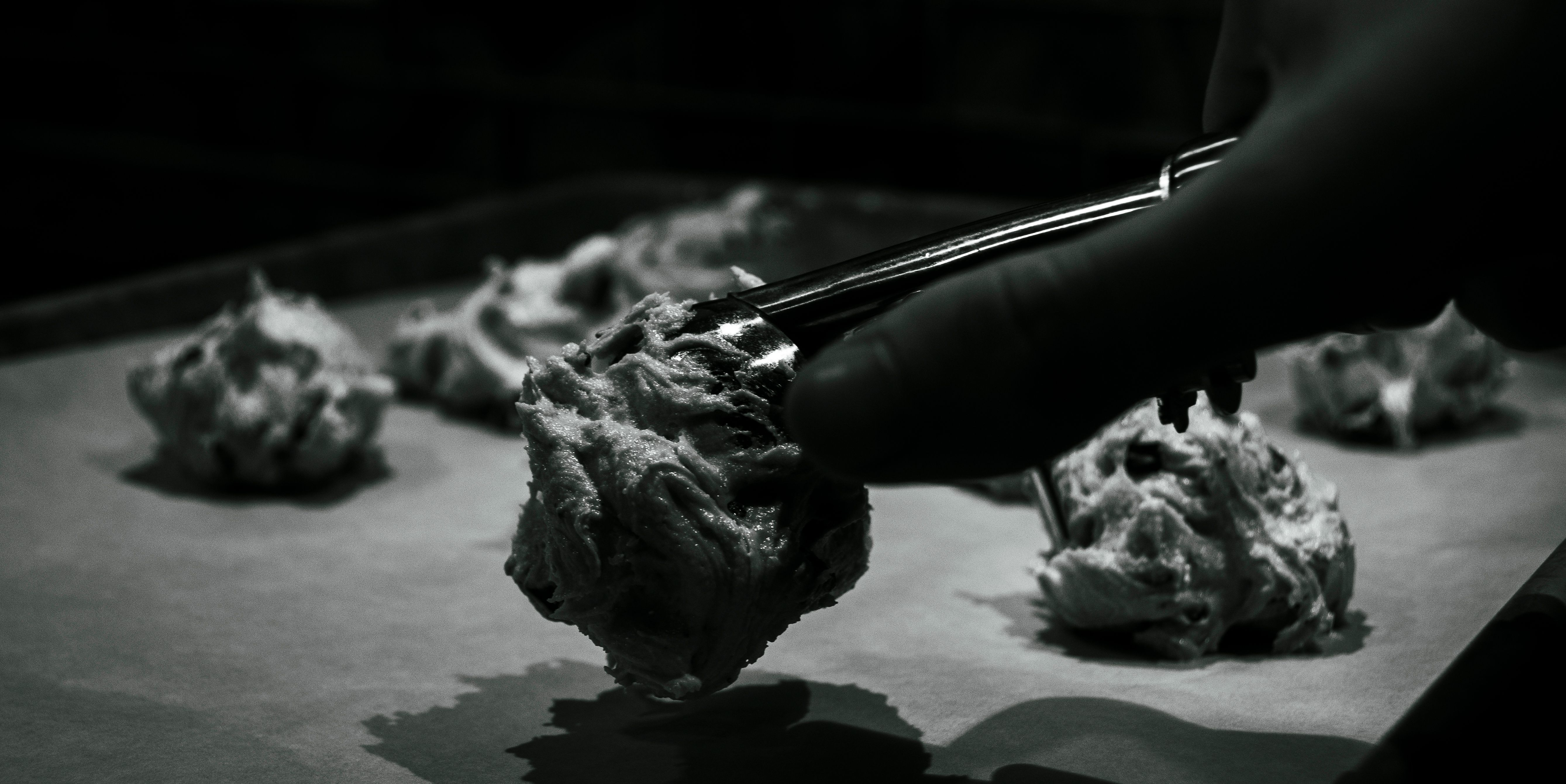 Black-and-white close-up of a gloved hand scooping cookie dough onto a baking sheet, dramatic side lighting