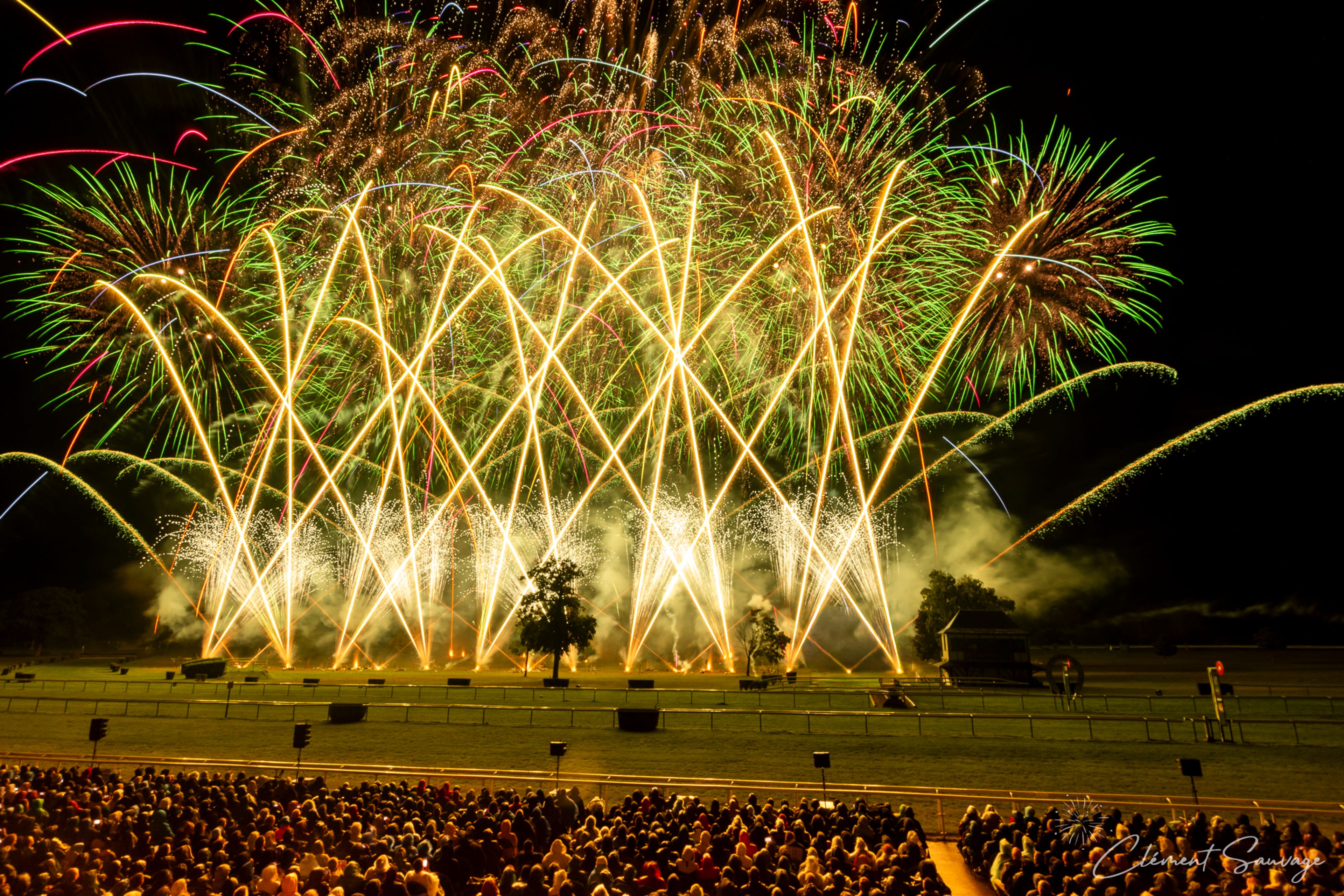 A large crowd watches a vibrant fireworks display with bright yellow arcs and bursts of green, red, blue, and gold lighting up the night sky over an open field in Compiègne, France, during the Masters de Feu 2025 event.