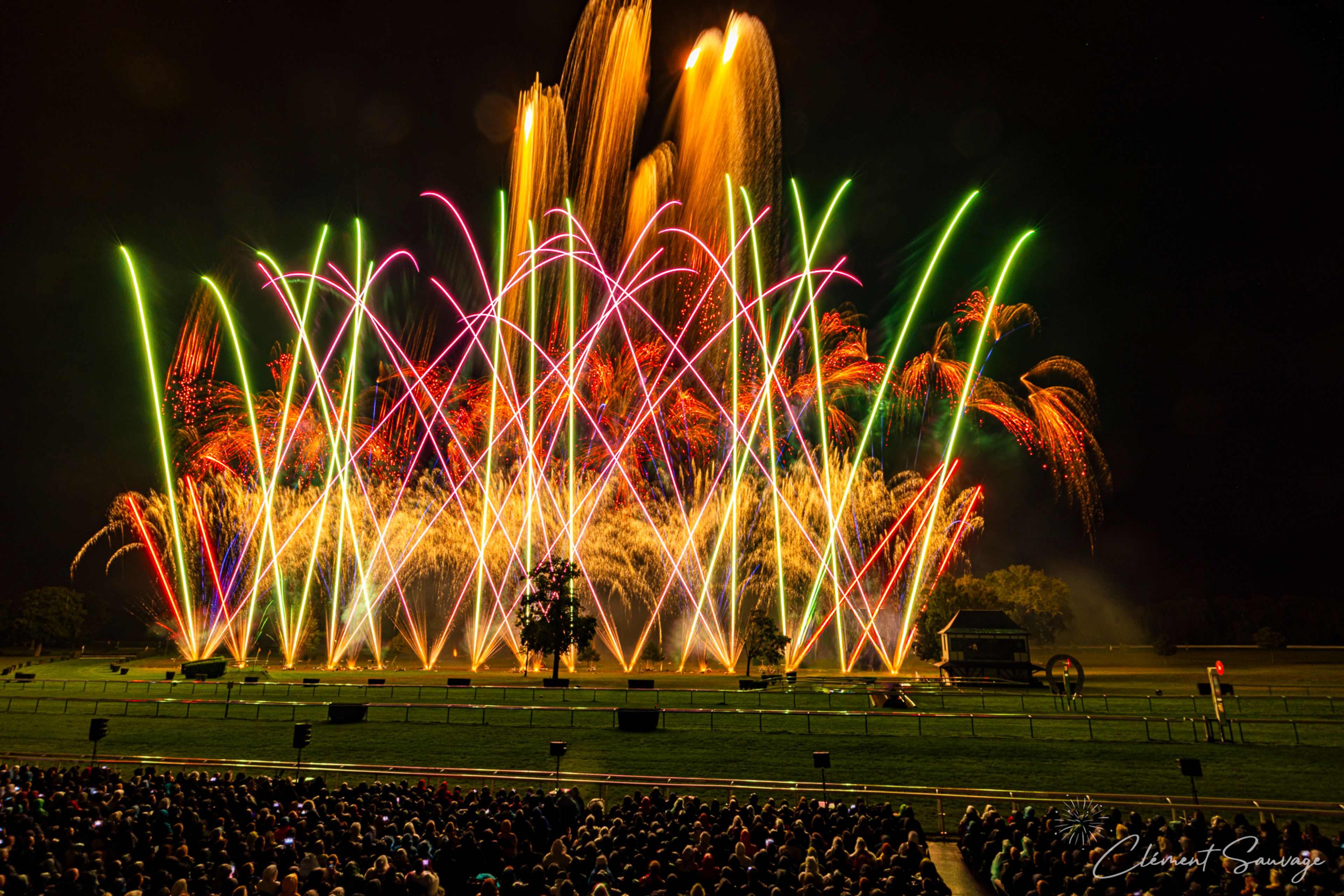 A large crowd watches a vibrant fireworks display with bright orange, green, red, and pink streams lighting up the night sky over a grassy field at the Masters de Feu event in Compiègne, France on September 20, 2025.