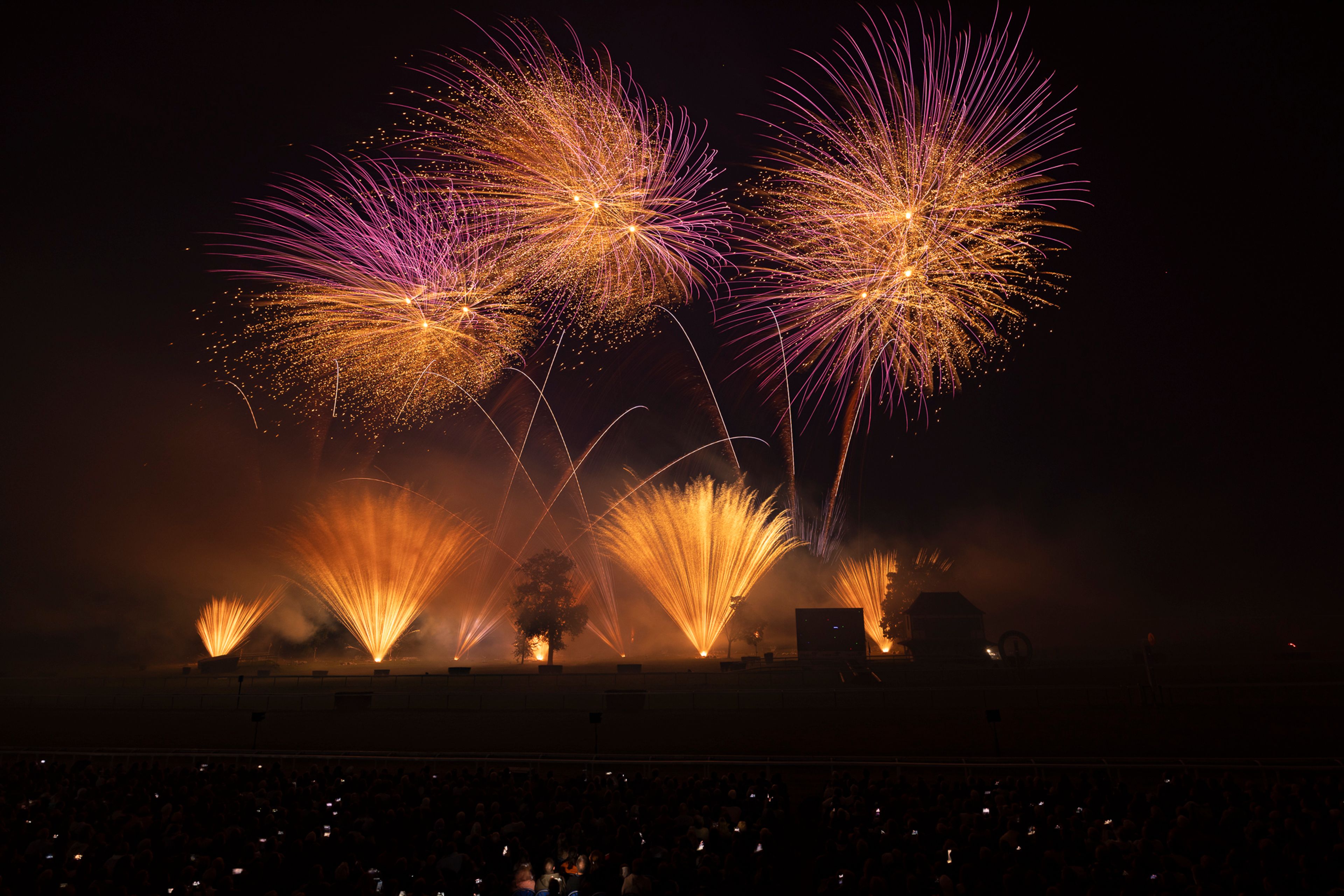 At Masters de Feu 2024 in Compiègne, France, a night sky is filled with large orange and purple fireworks bursts above a dark landscape with trees and structures, while an audience watches from below, some holding up phone lights.
