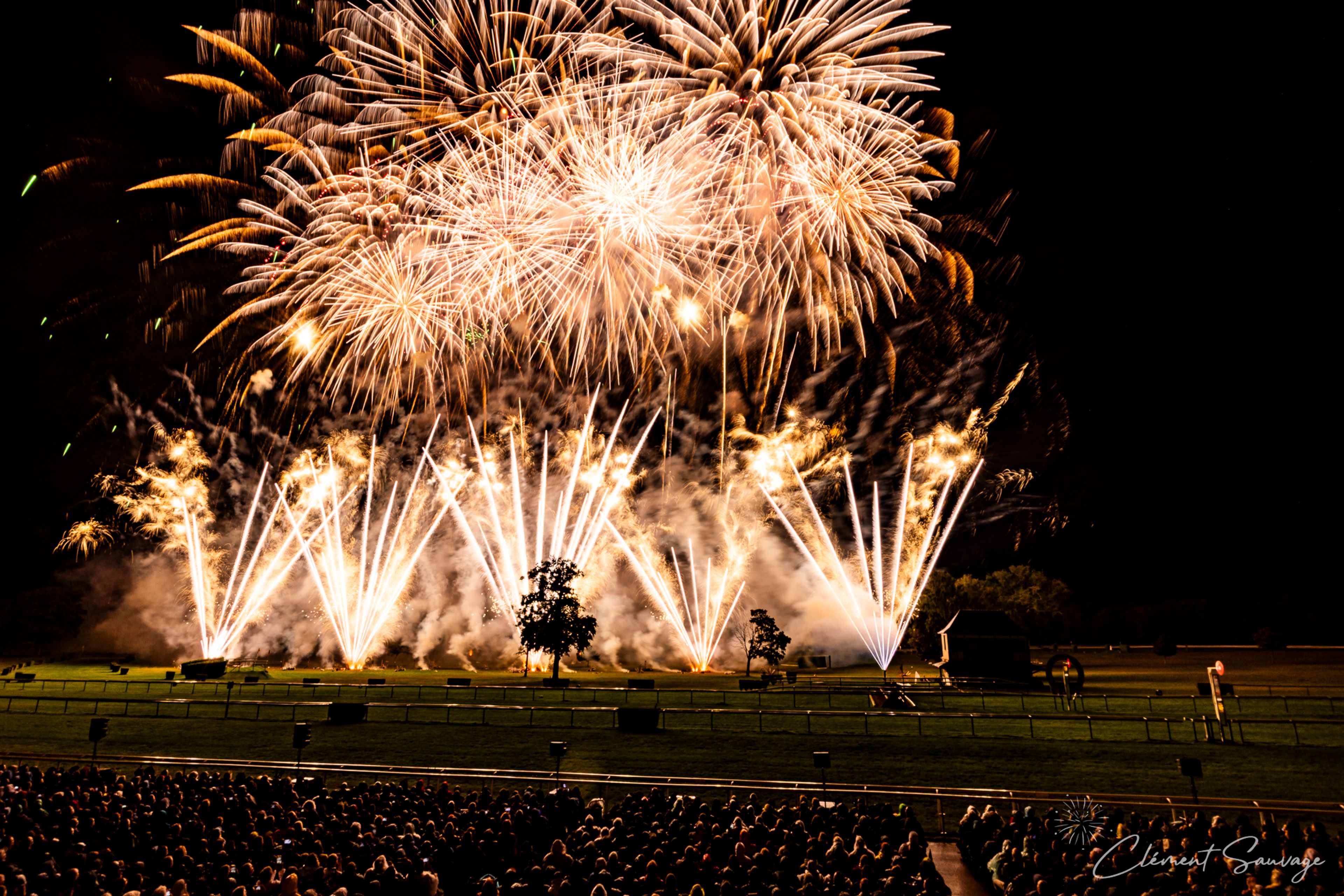 A large crowd watches a fireworks display with numerous golden and white explosions lighting up the night sky over a grassy field with scattered trees, during the Masters de Feu event in Compiègne, France, on September 20, 2025.