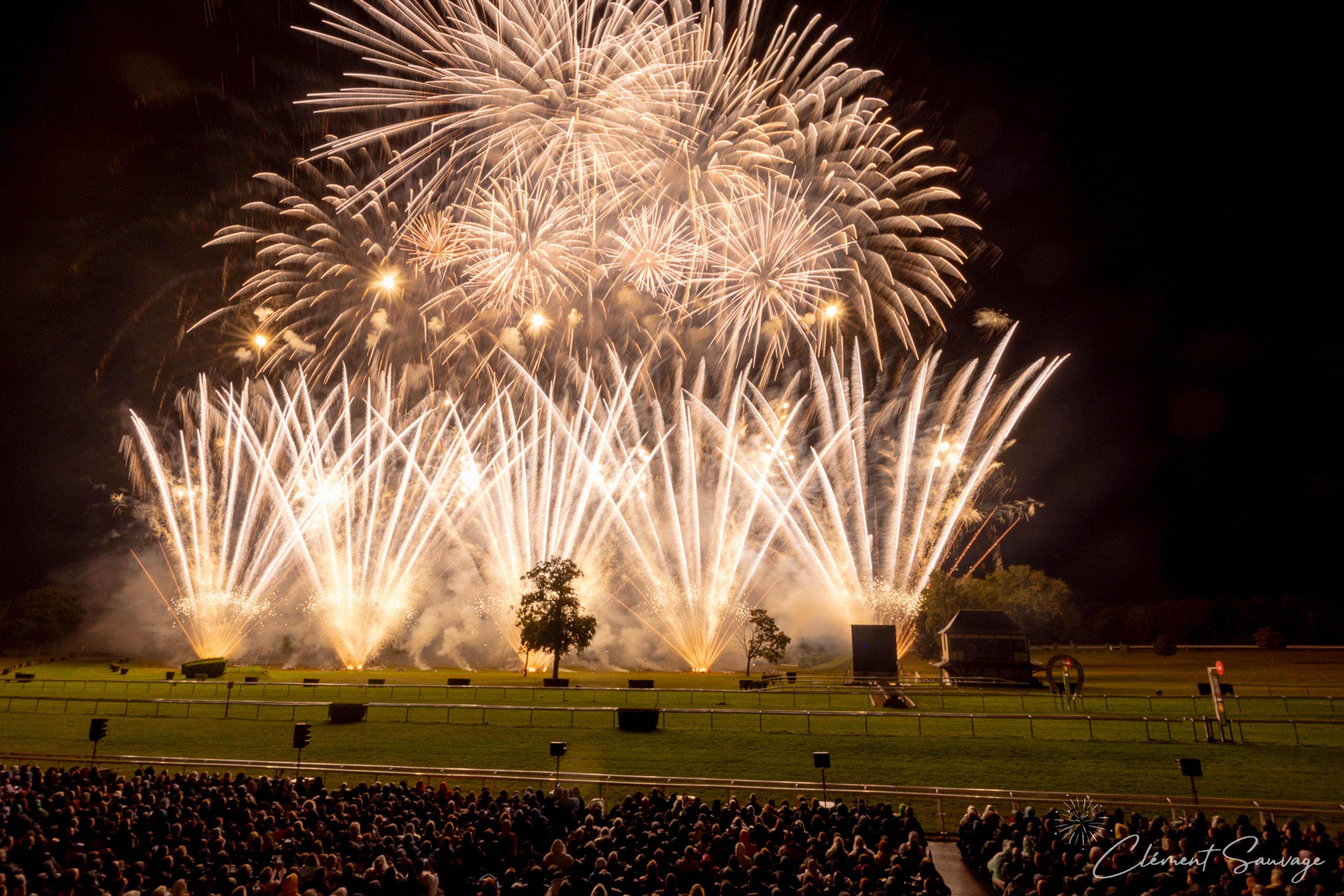 A large crowd watches a fireworks display at the Masters de Feu 2025 event in Compiègne, France, with bright white and gold fireworks bursting and fanning out in the night sky above a grassy field with a few trees and structures.