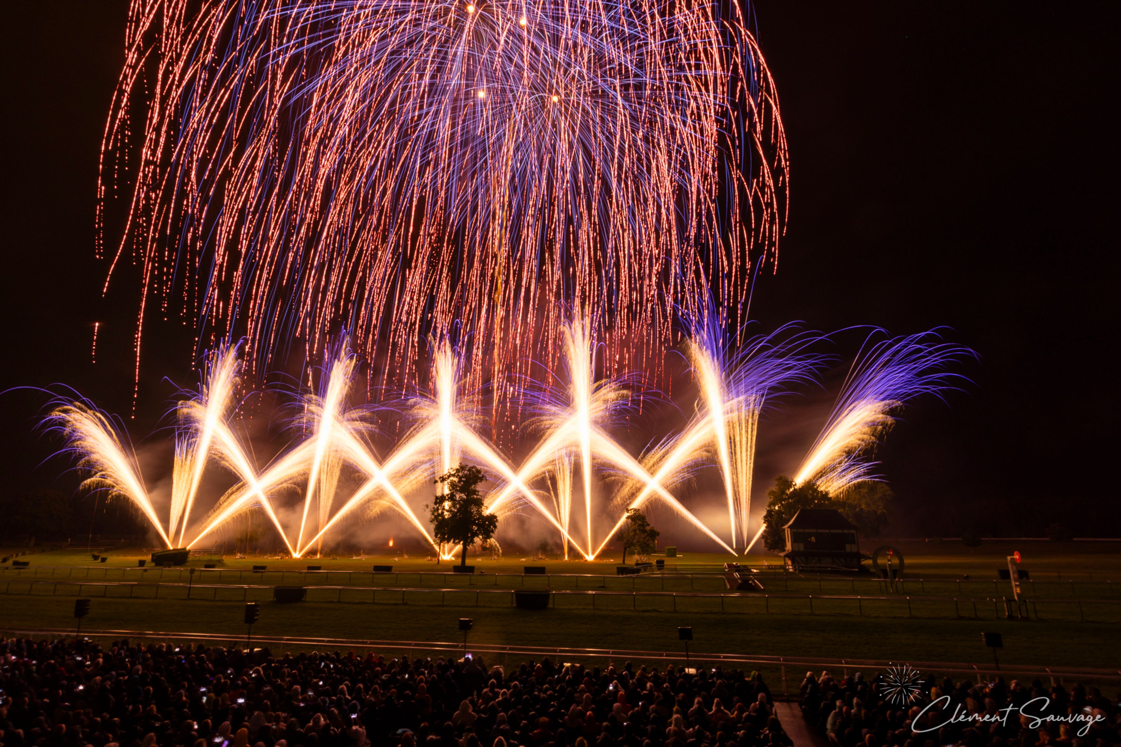 At the Masters de Feu 2025 in Compiègne, France, fans watch a nighttime fireworks display featuring tall orange and purple pyrotechnic fountains exploding in the sky above a green field with scattered trees and a small wooden building.