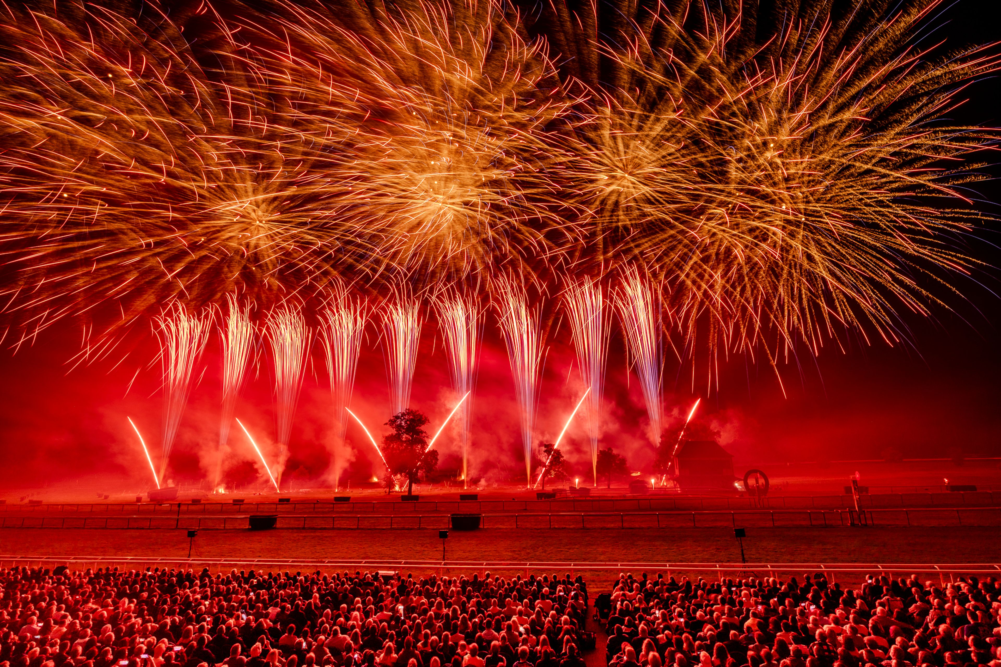 A large crowd watches a fireworks display at night in Compiègne, France, during Masters de Feu 2024, with golden and red fireworks bursting and cascading over a silhouetted landscape illuminated by red light and smoke.