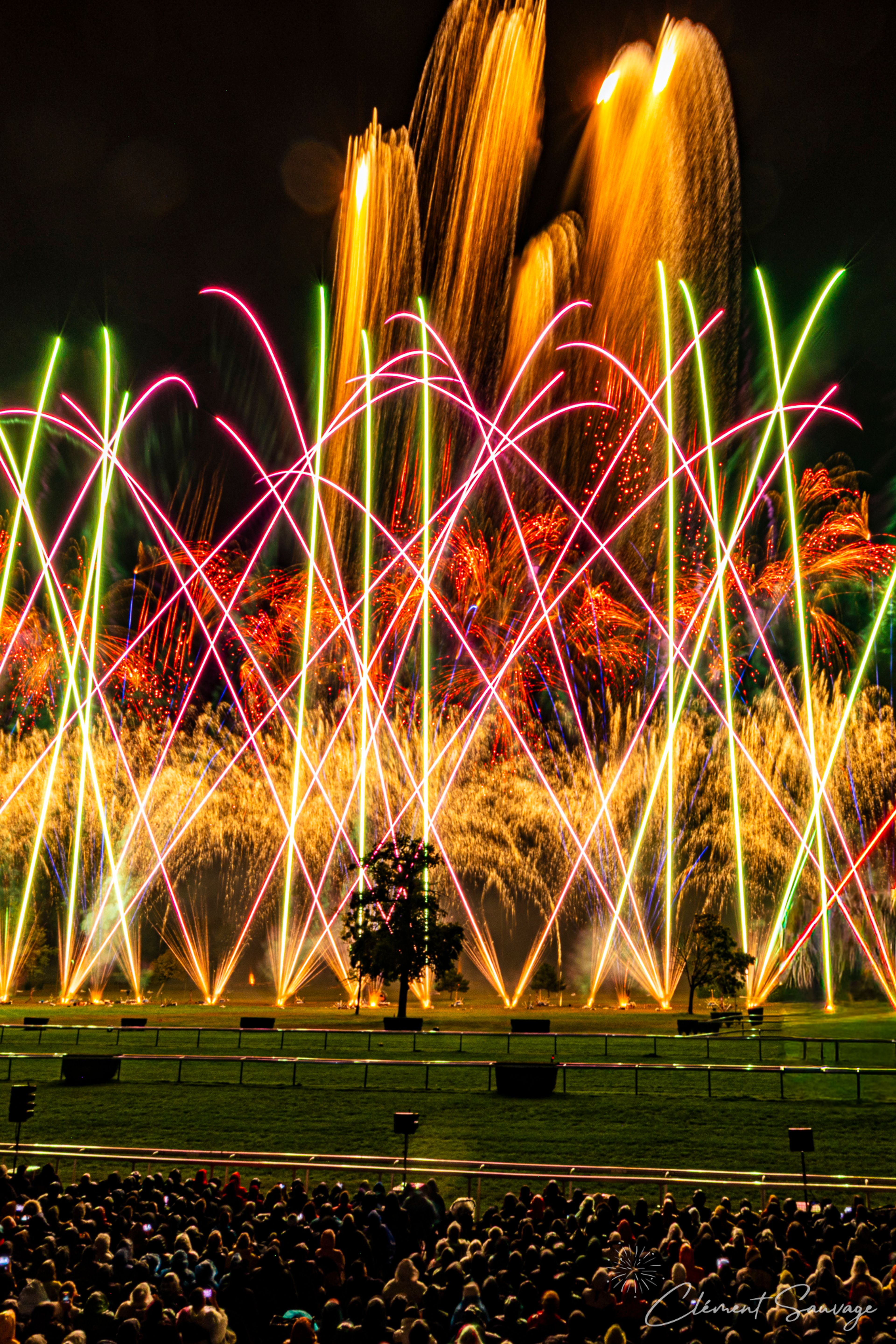 A nighttime fireworks display at Masters de Feu 2025 in Compiègne, France, shows bright orange, red, green, and pink arcs and sparks lighting up the sky above a grassy field with silhouetted trees and an audience in the foreground.