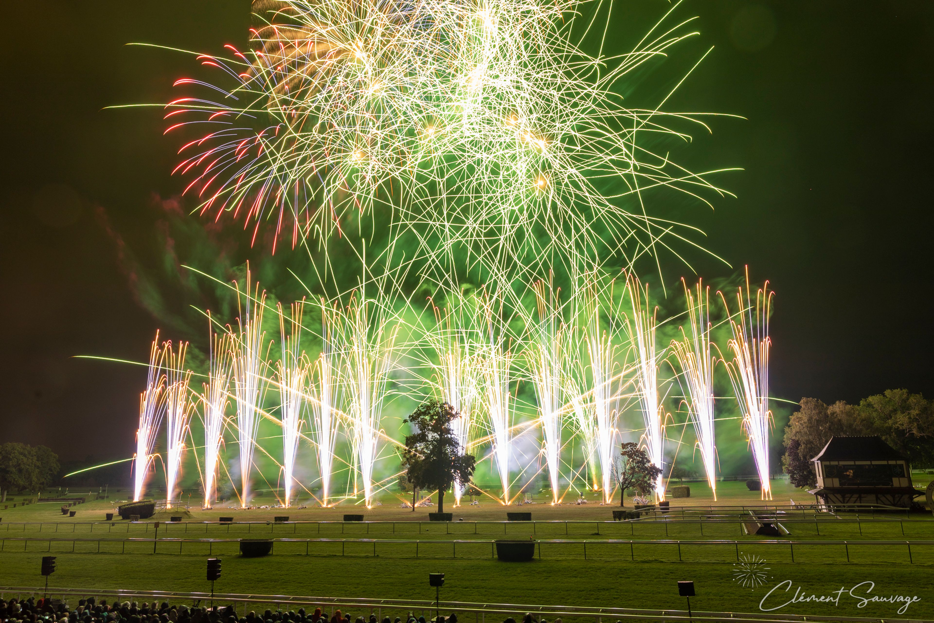 Bright green, white, red, and yellow fireworks light up the night sky over a grassy field with scattered trees and a small wooden structure, while a crowd watches from the foreground at the Masters de Feu event in Compiègne, France on September 20, 2025.