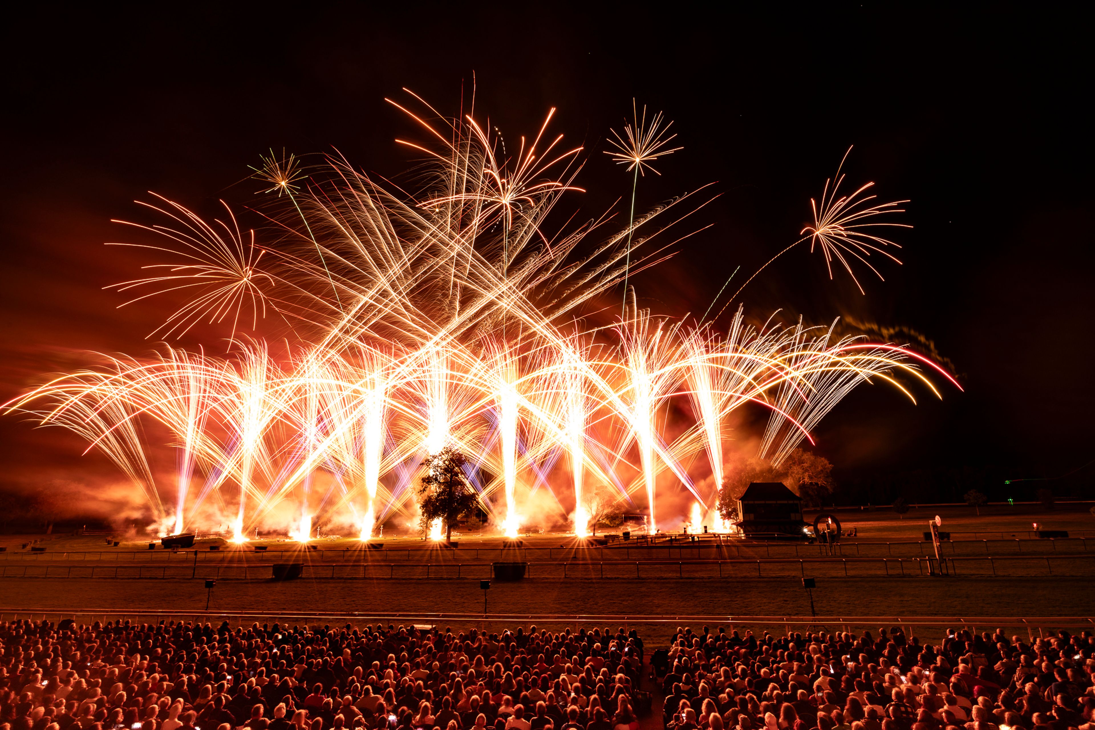 A large crowd watches an elaborate fireworks display at night during Masters de Feu 2024 in Compiègne, France, with bright orange, white, and red sparks shooting in various directions against the dark sky.