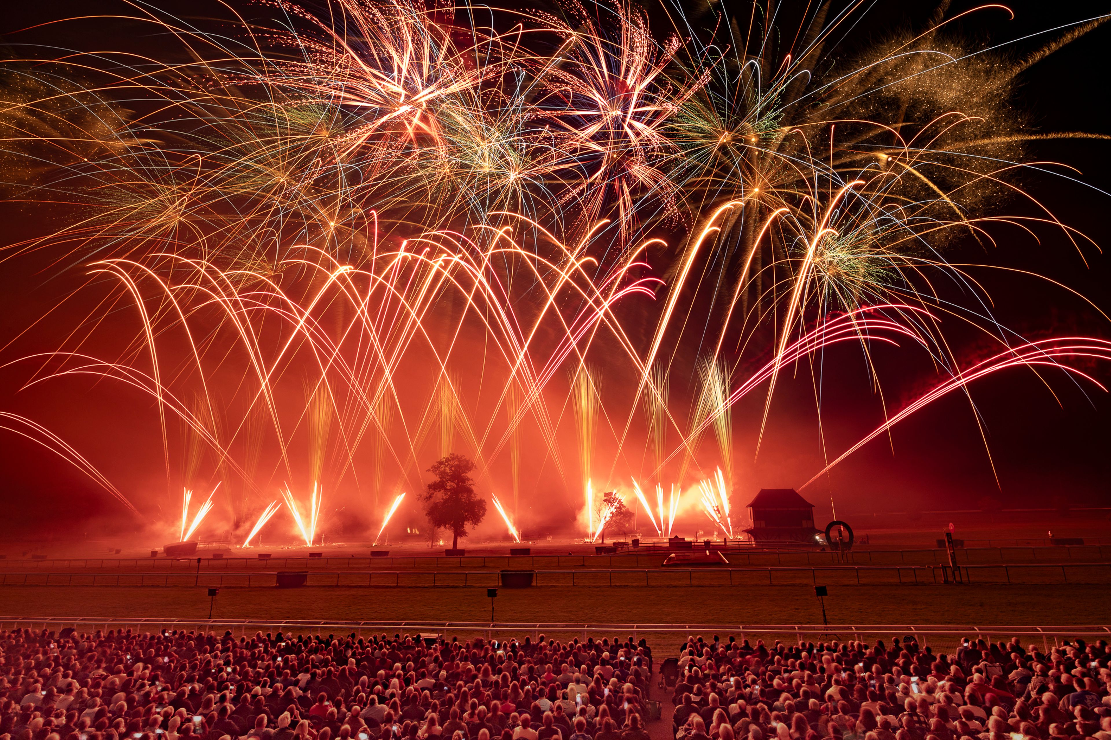 A large crowd watches a vibrant fireworks display featuring red, orange, yellow, and green bursts over a dark field with a single tree and a small building during the Masters de Feu 2024 event in Compiègne, France.