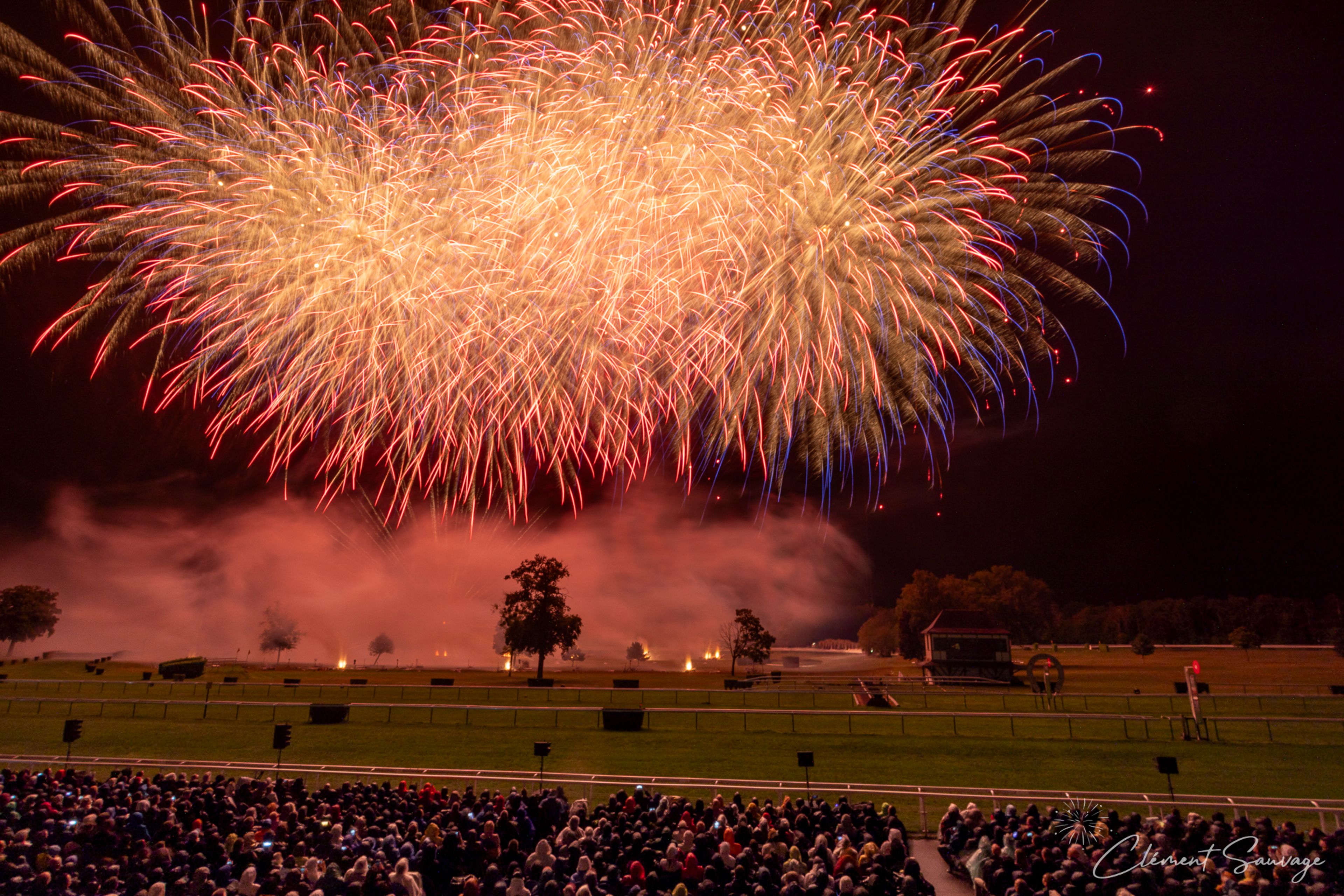 A large crowd watches a vibrant fireworks display with golden, red, and blue streaks lighting up the night sky above a grassy field with scattered trees during the Masters de Feu 2025 event in Compiègne, France.