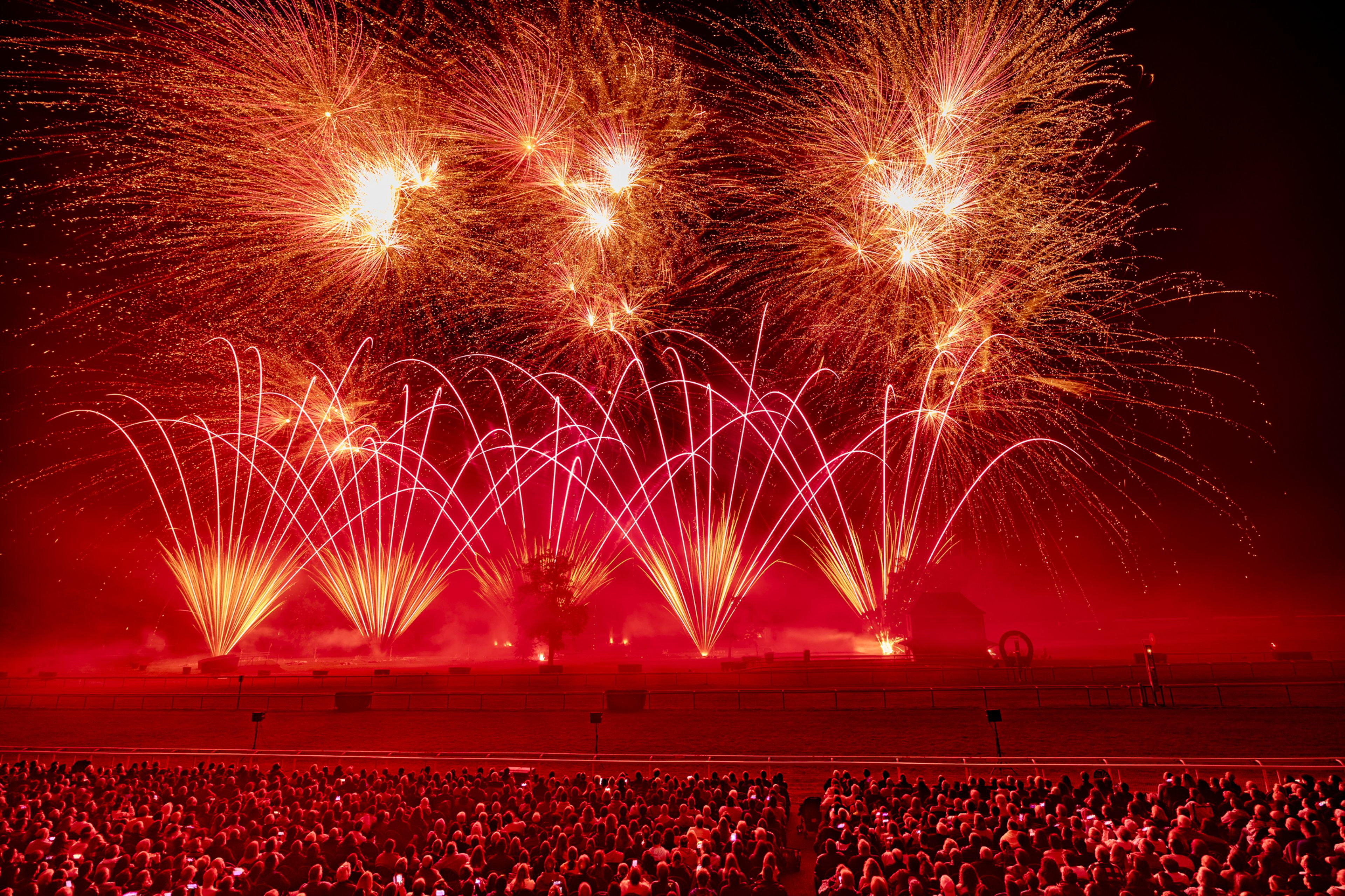 A large crowd watches a vibrant display of red and orange fireworks lighting up the night sky at the Masters de Feu 2024 event in Compiègne, France, with multiple bursts and arcs of sparks illuminating the scene.